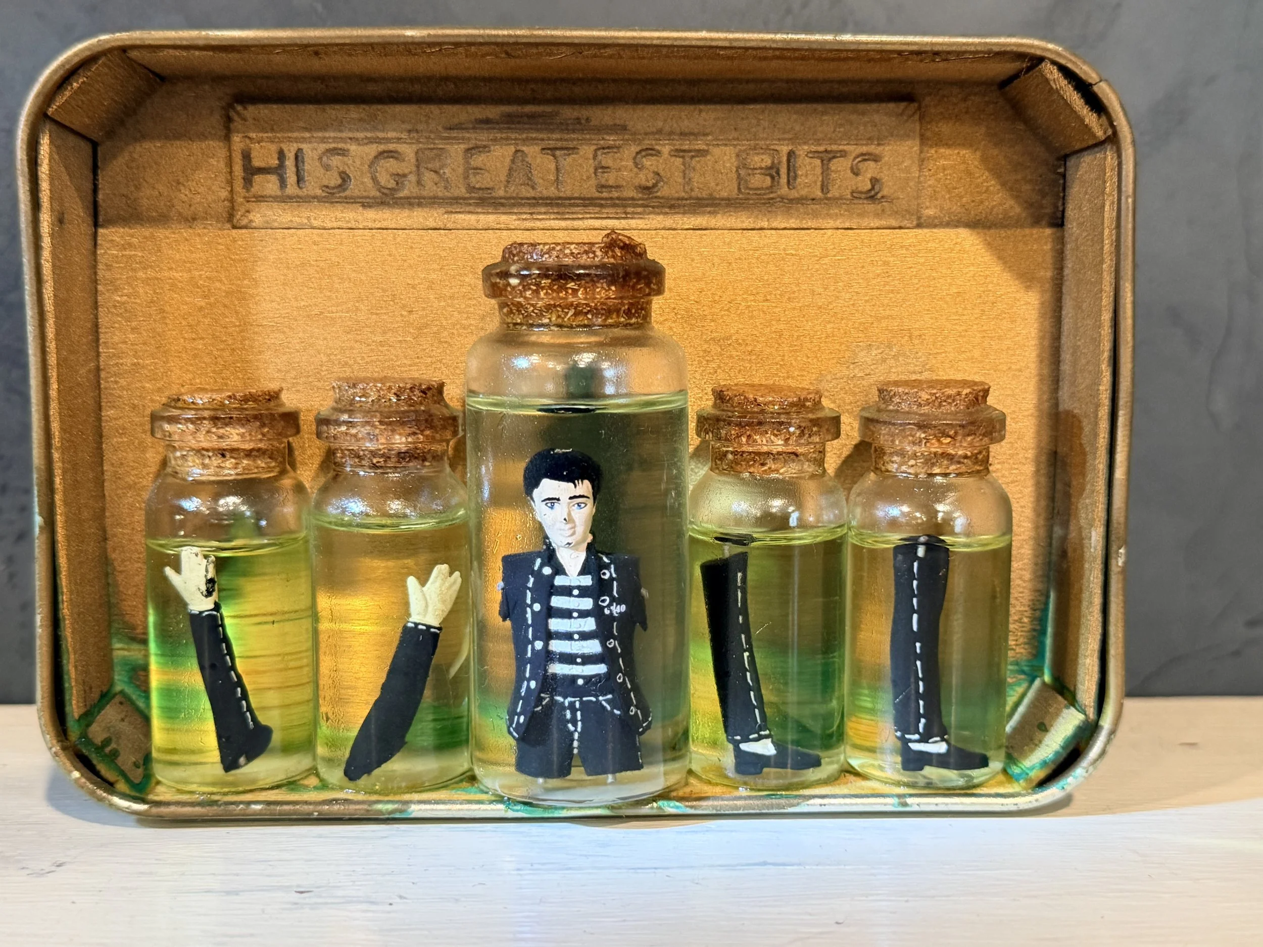 A small glass vial with a figurine of a man in a black suit and striped shirt inside, placed inside a vintage-style jewelry box with the inscription 'HIS GREATEST BITS' engraved inside.
