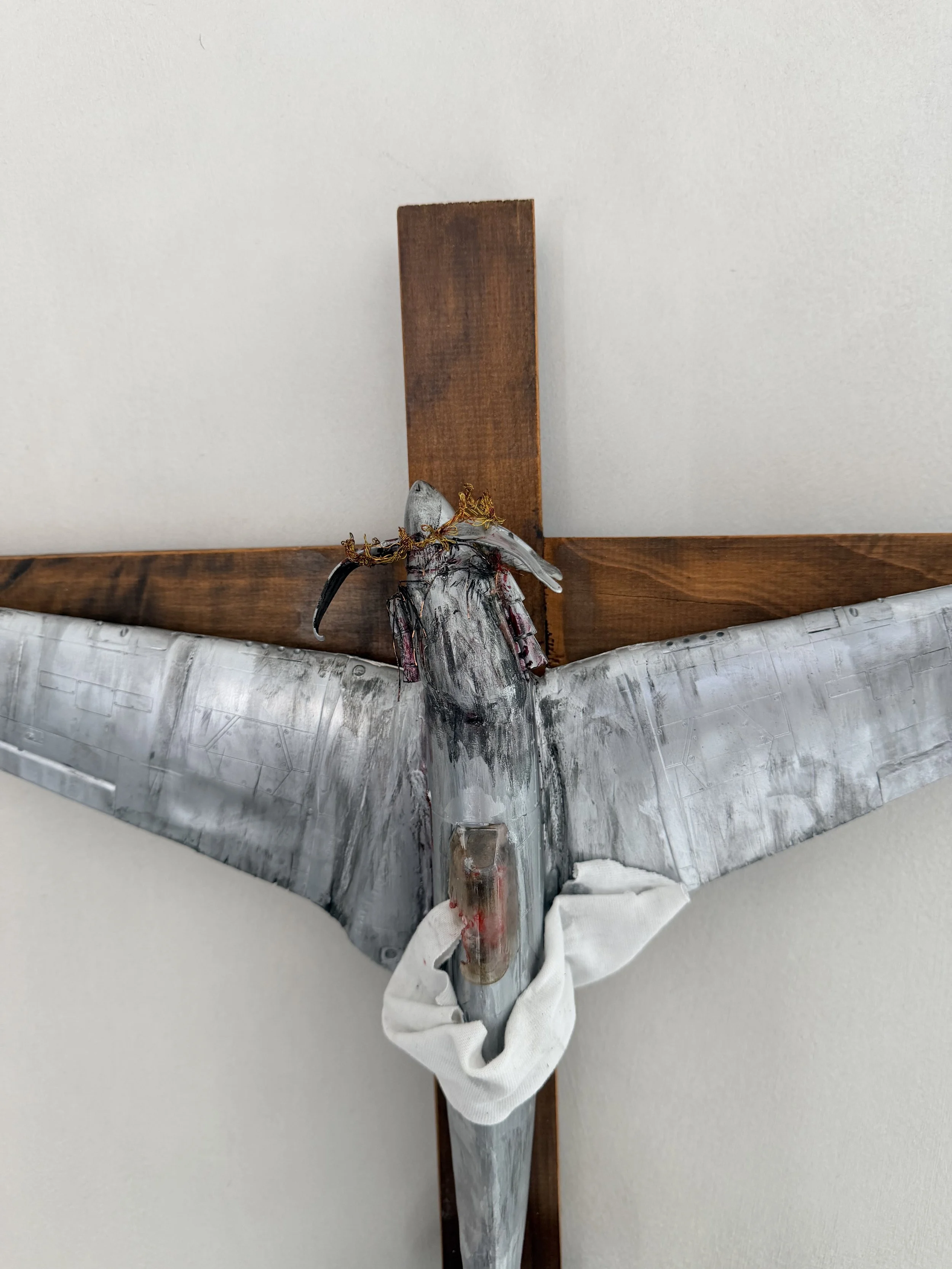 A metallic sculpture of a bird mounted on a wooden cross, decorated with a gold wire crown, with a white cloth draped below.