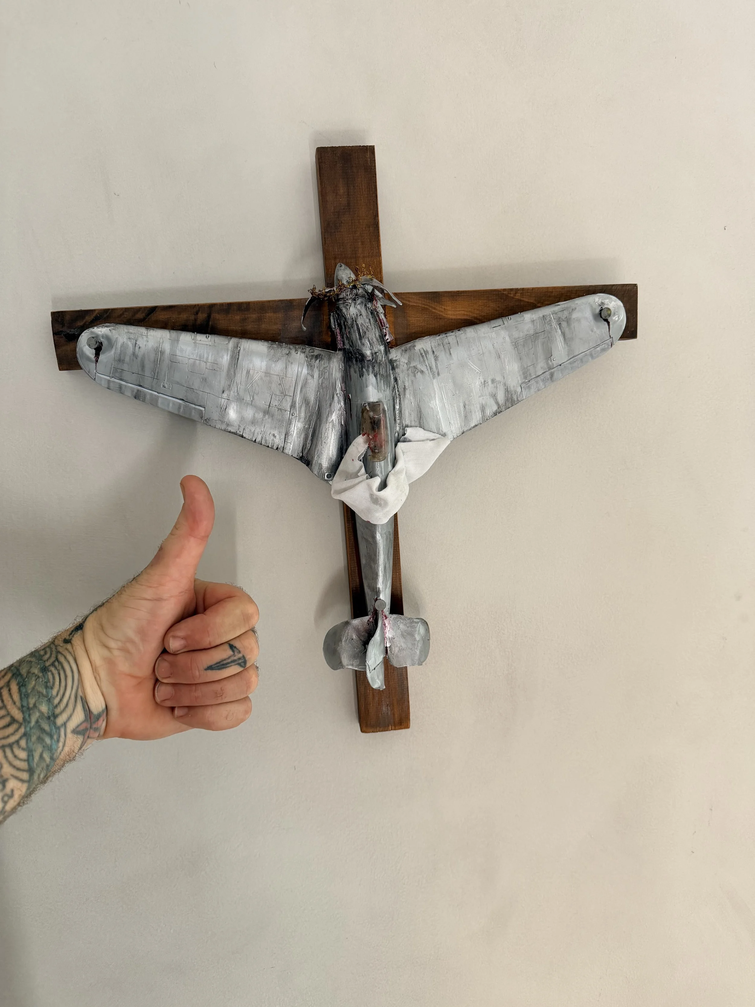 A tattooed hand pointing at a metallic model of a flying bird mounted on a wooden cross.