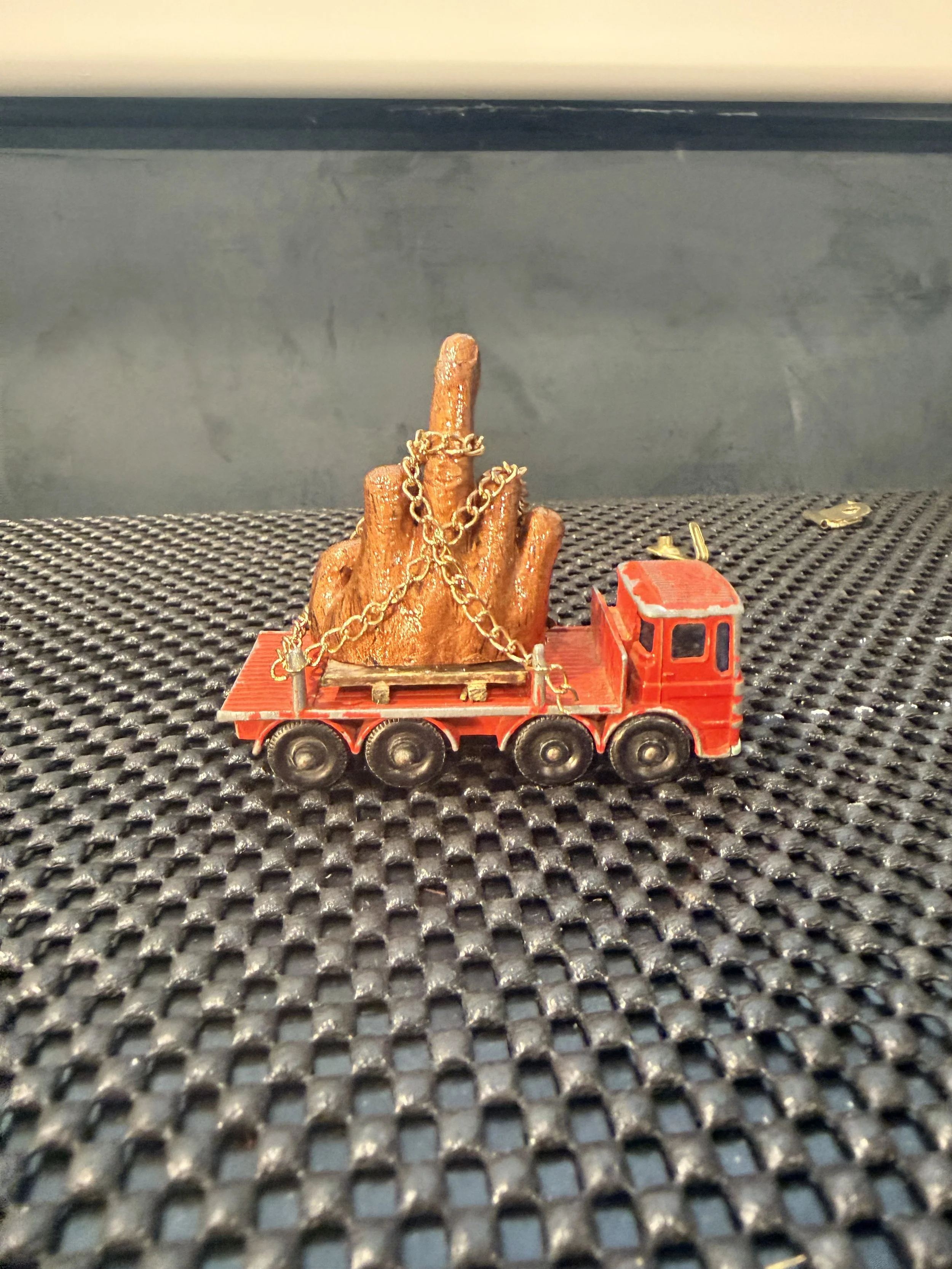 A small toy red semi-truck carrying a large wooden sculpture of a hand with gold chains wrapped around it, placed on a black textured surface.