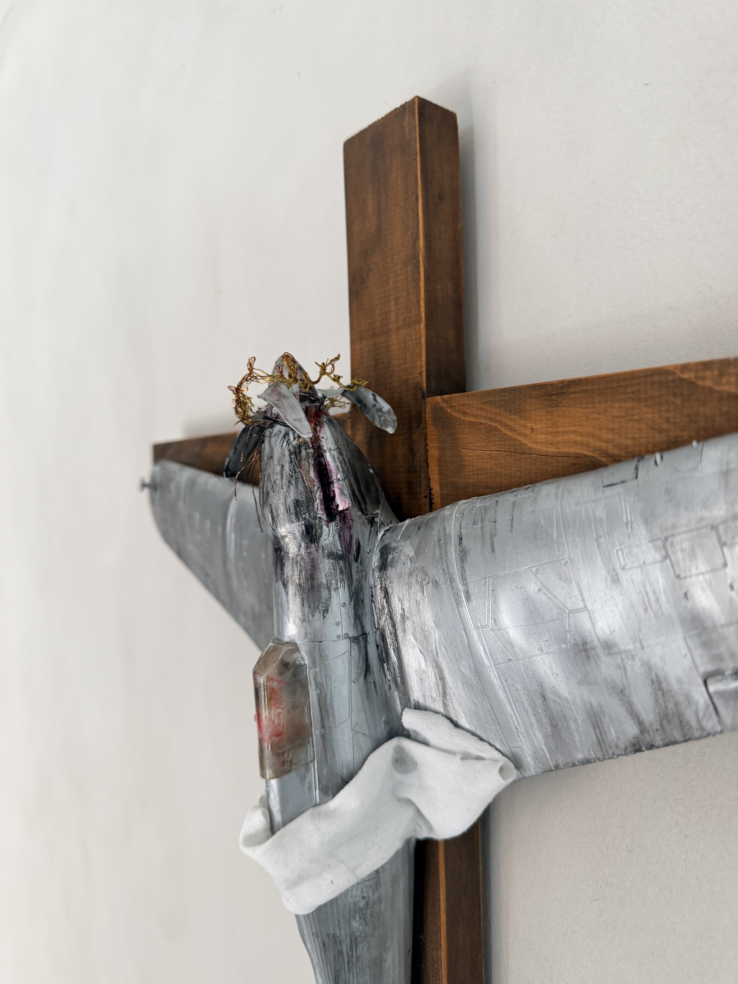 A metallic fire alarm with a silver tube and a copper-colored component resting on a white cloth on a flat surface, with a wooden block behind it and a plain white wall in the background.
