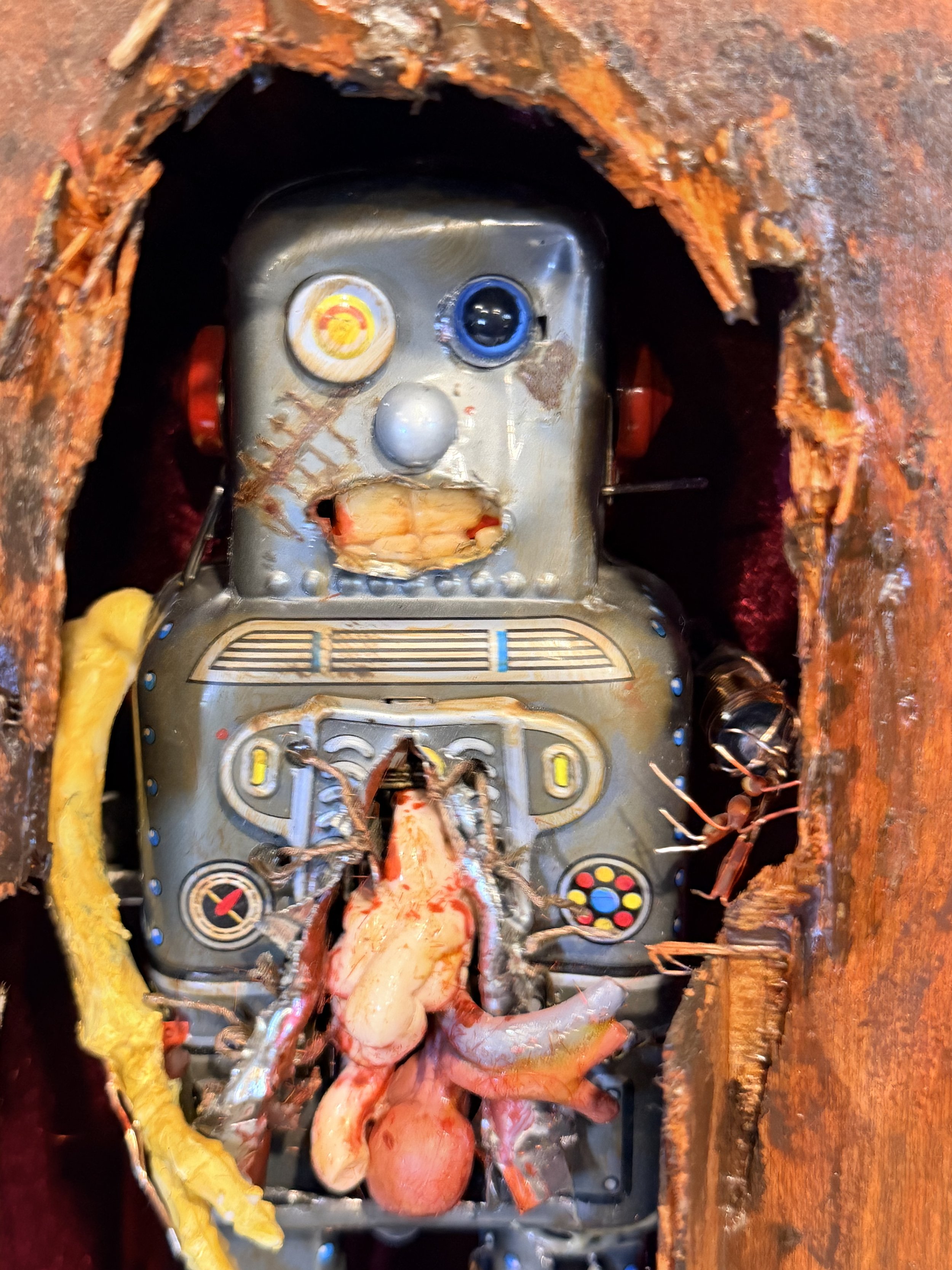 A robotic device with a human-like face, featuring a yellow eye and a blue eye, encased in a metal casing with various buttons and dials, surrounded by wires and meat, and embedded in a rusty, corroded surface.
