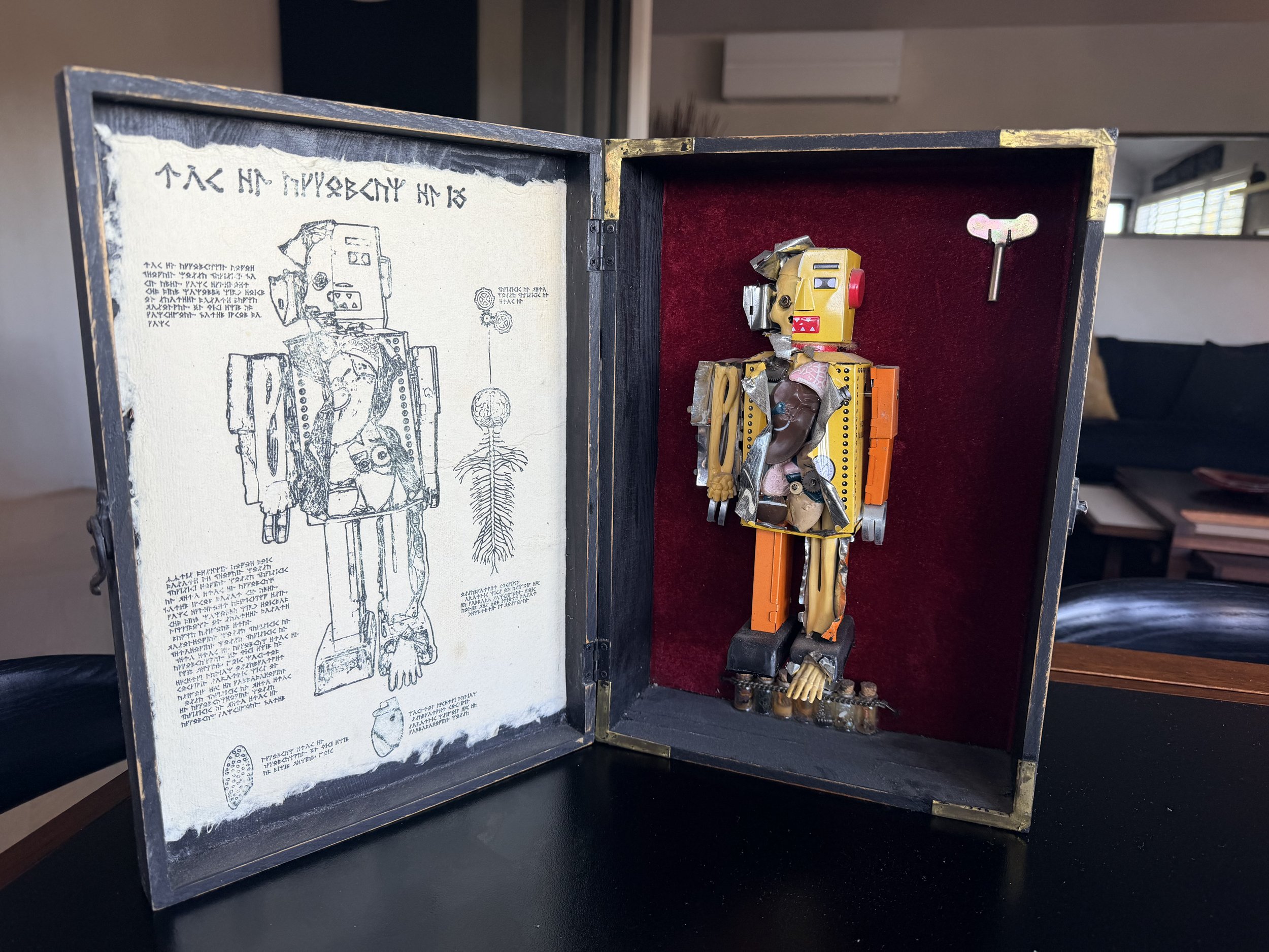 A decorated box featuring a vintage robot figure made of various mechanical parts, set against a red velvet background. The interior of the box shows an illustrated paper with handwritten text and drawings, including a human skeleton and other scient