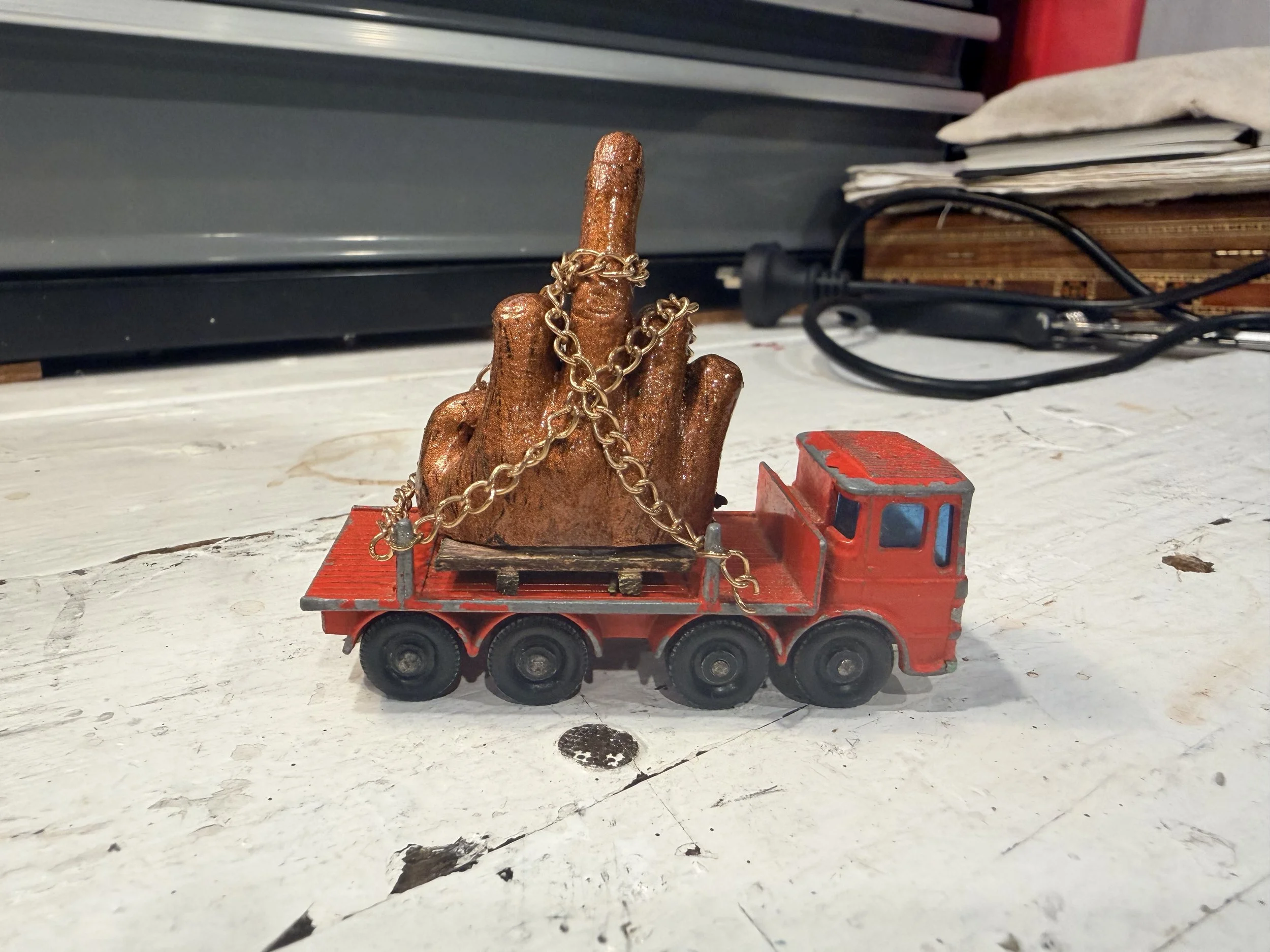 A small toy train with a red caboose is carrying a large, glittery, bronze-colored hand sculpture with an upraised middle finger. The sculpture is decorated with multiple gold chains. The scene is set on a white, worn table with visible scratches and