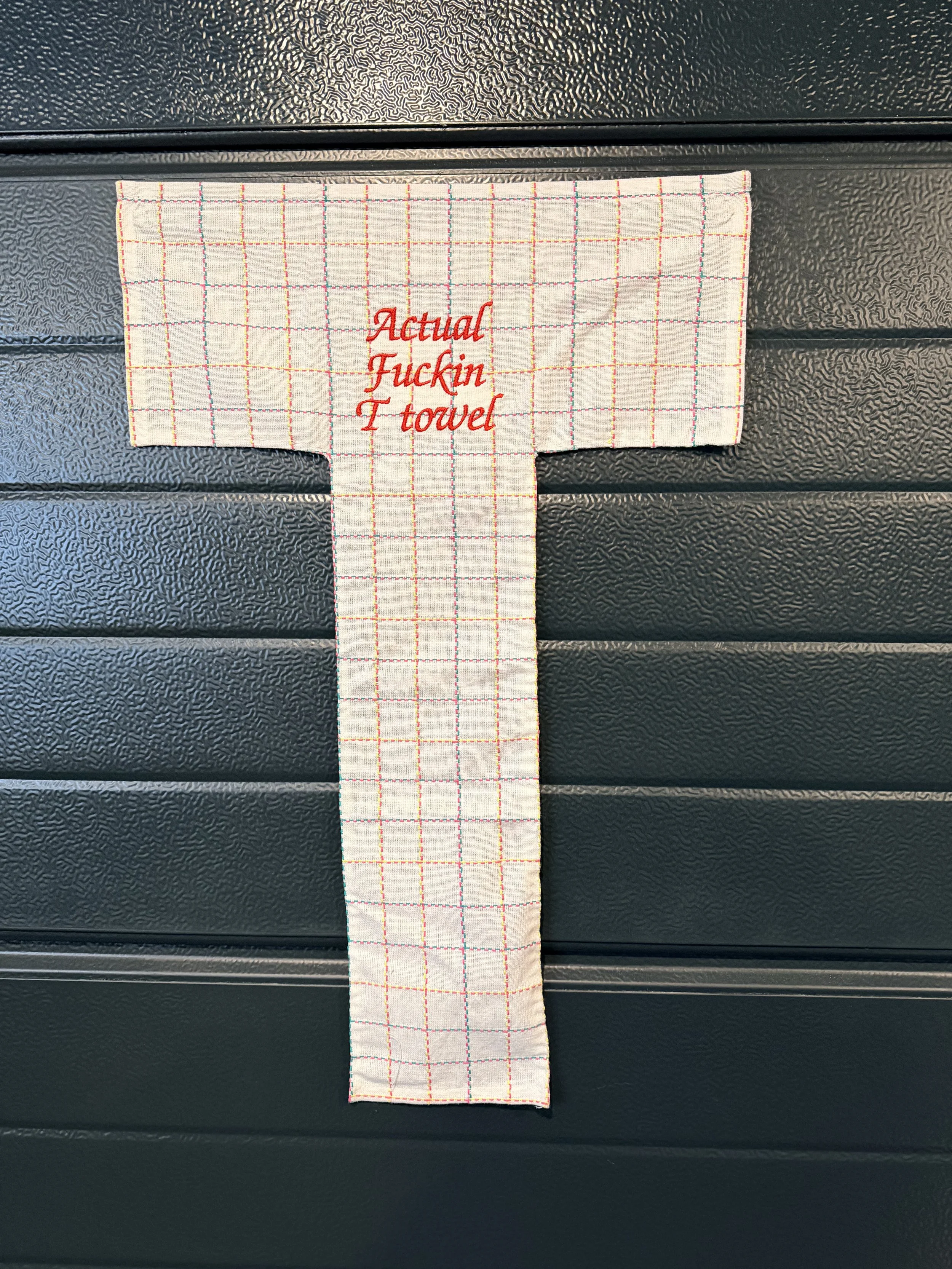 A white dish towel with multicolored grid lines and red embroidered text that reads "Actual F***in Towel," hanging on a black textured surface.