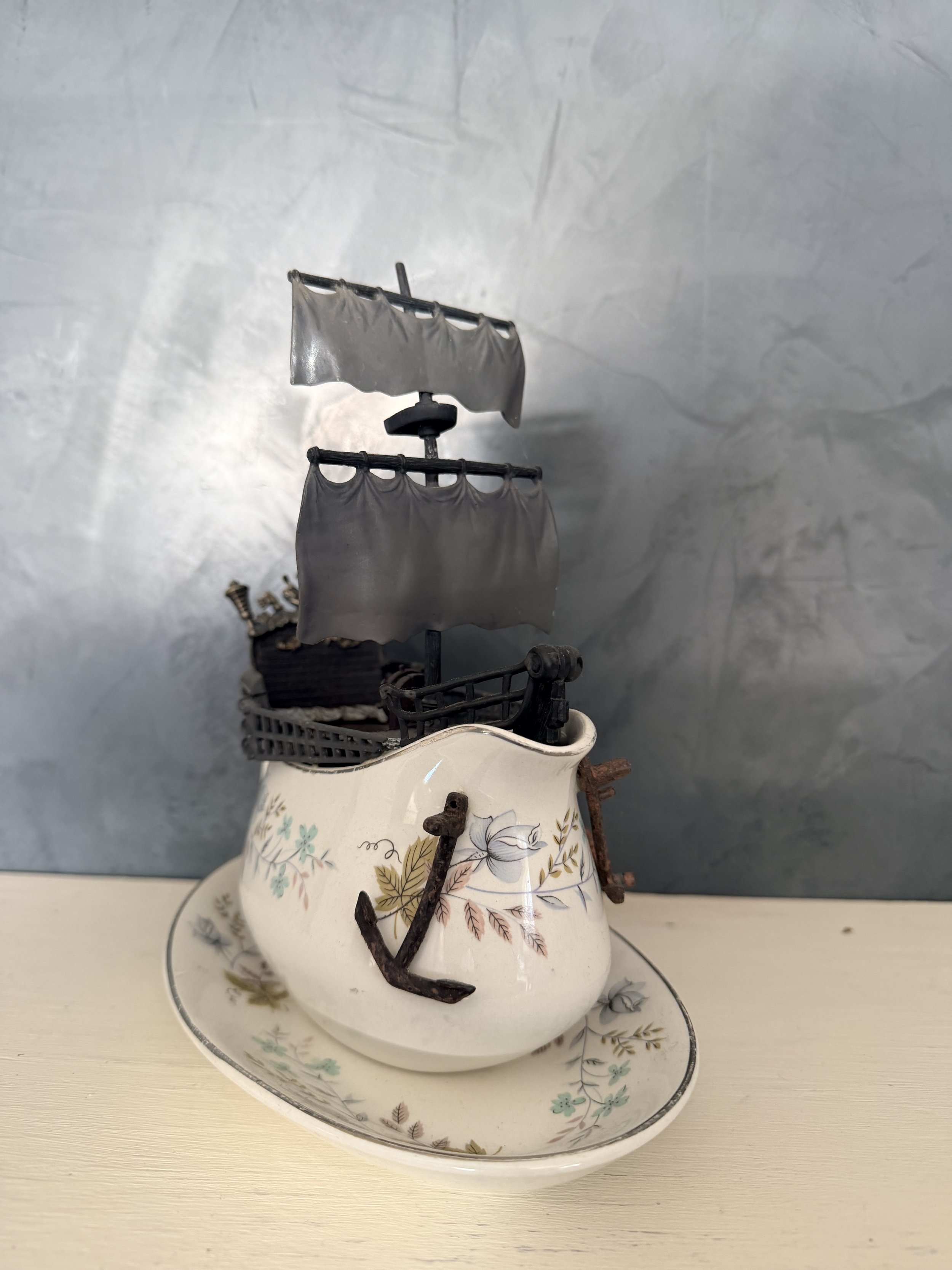 A vintage teapot with floral decoration holding a small ship model with fabric sails, placed on small matching dishes and a plate.