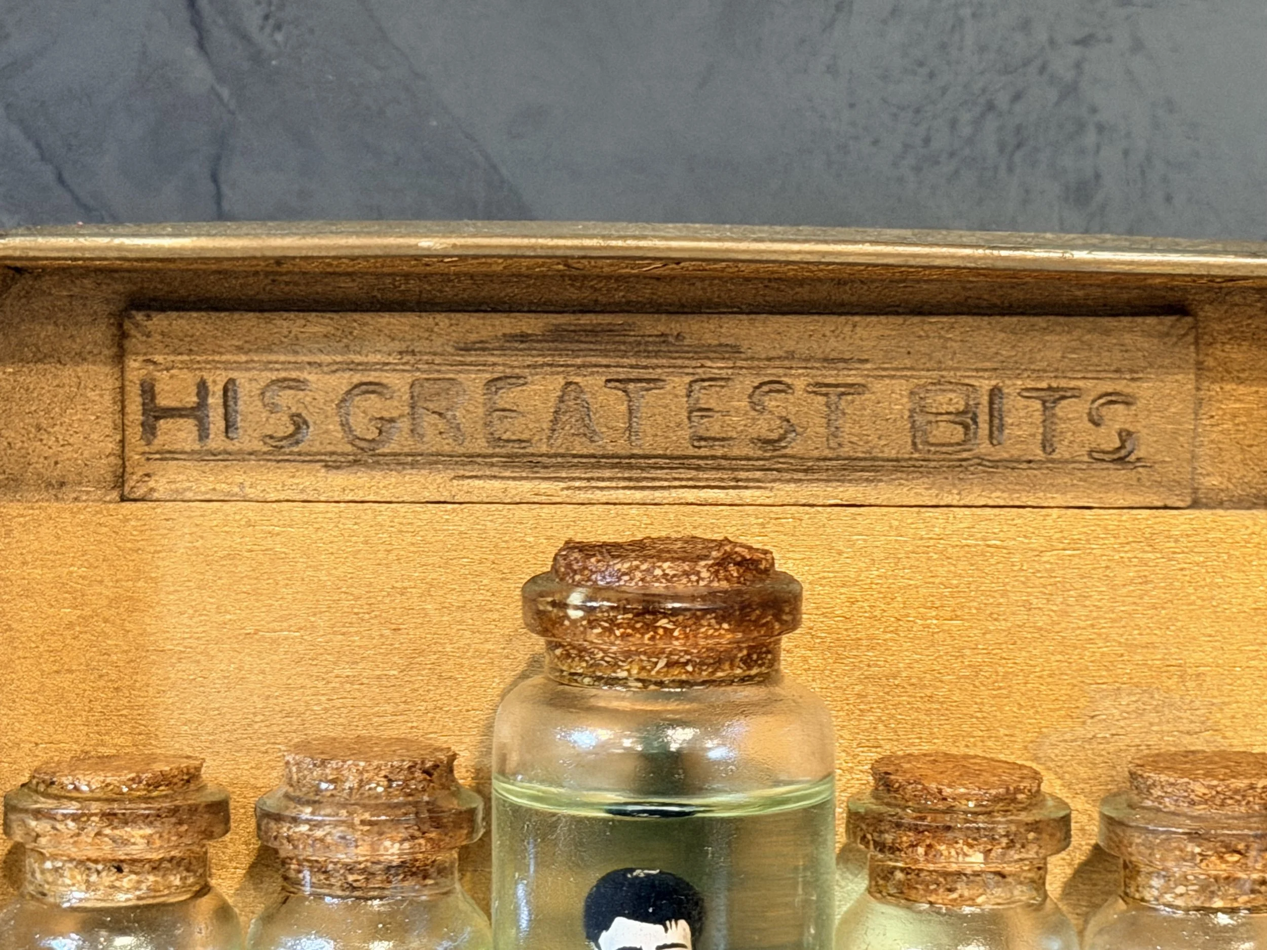 Close-up of small glass bottles with cork stoppers, some with handwritten labels, on a wooden surface, behind a wooden box with the word 'HIS' engraved on it, against a textured background.