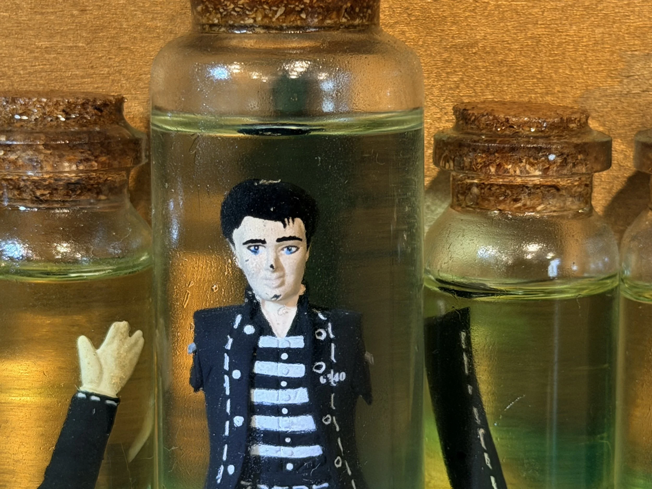 Close-up of small glass bottles with cork stoppers, with one featuring a painted portrait of a woman with short dark hair, blue eyes, and a black jacket with white and black striped shirt underneath, set against a wooden surface background.