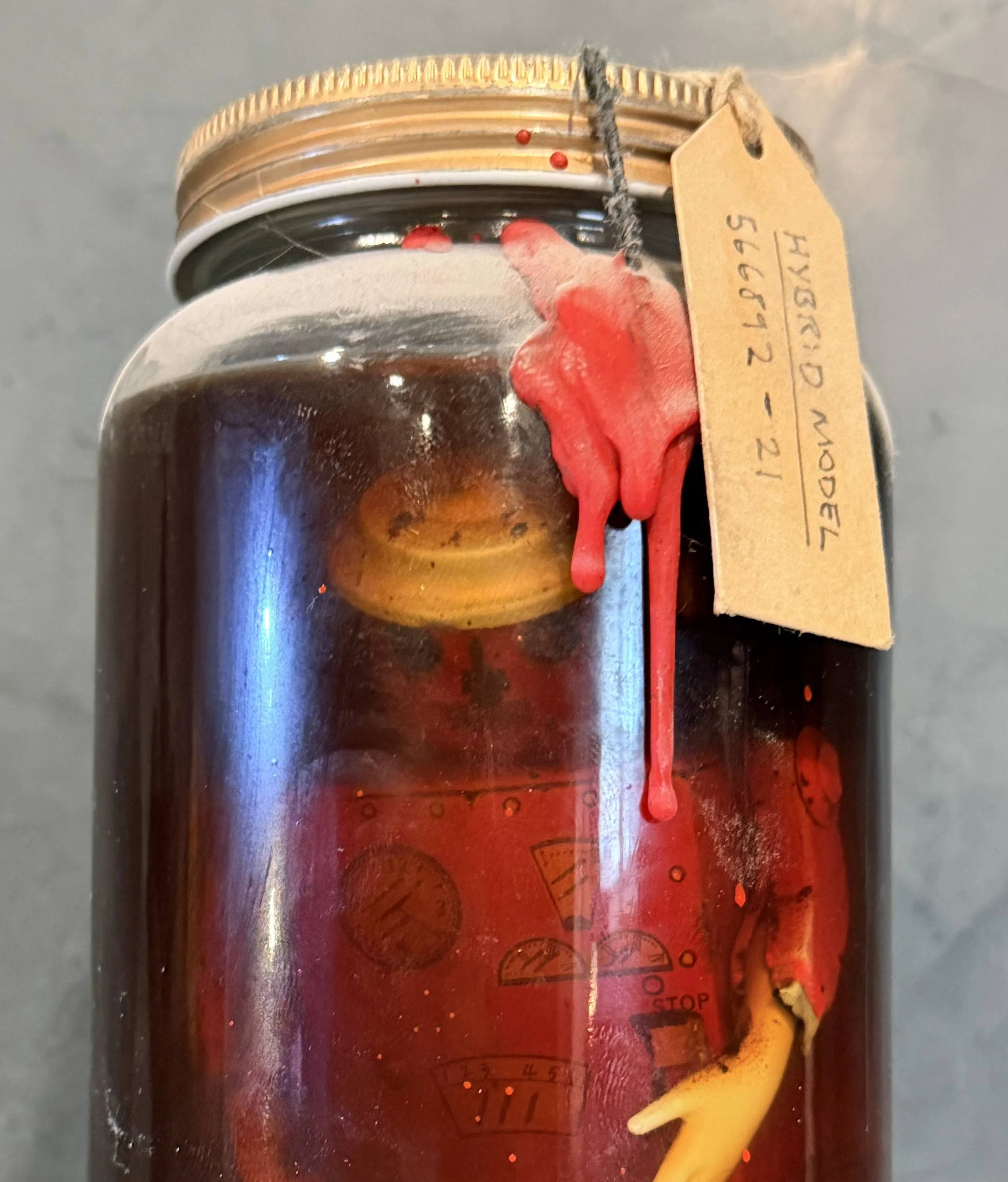 A glass jar filled with a dark liquid with a piece of banana and red substance dripping down the side, and a warning tag attached to the jar.
