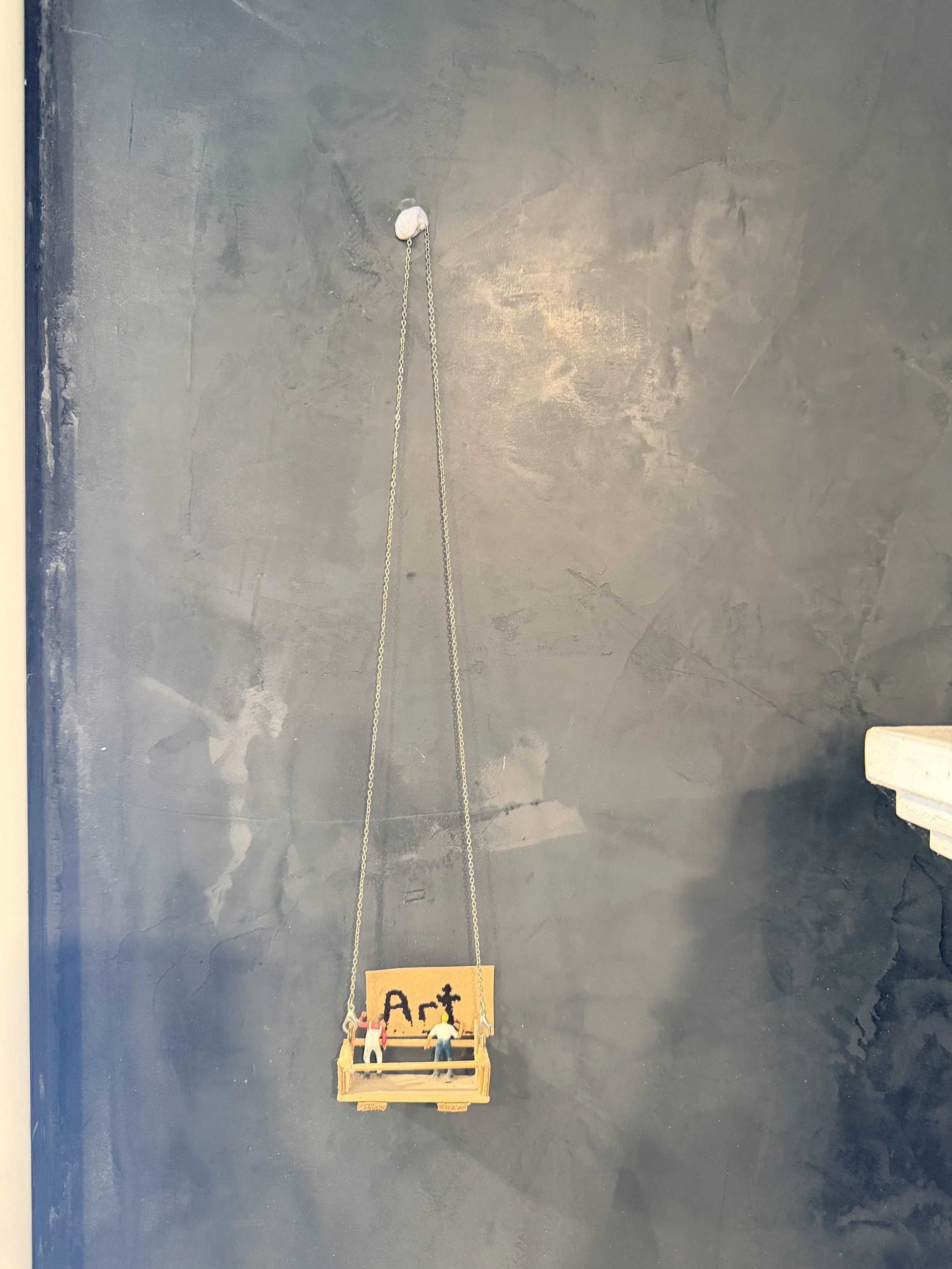A small wooden art display labeled "Art" hanging by a chain on a dark wall. The display holds four miniature figurines of people.
