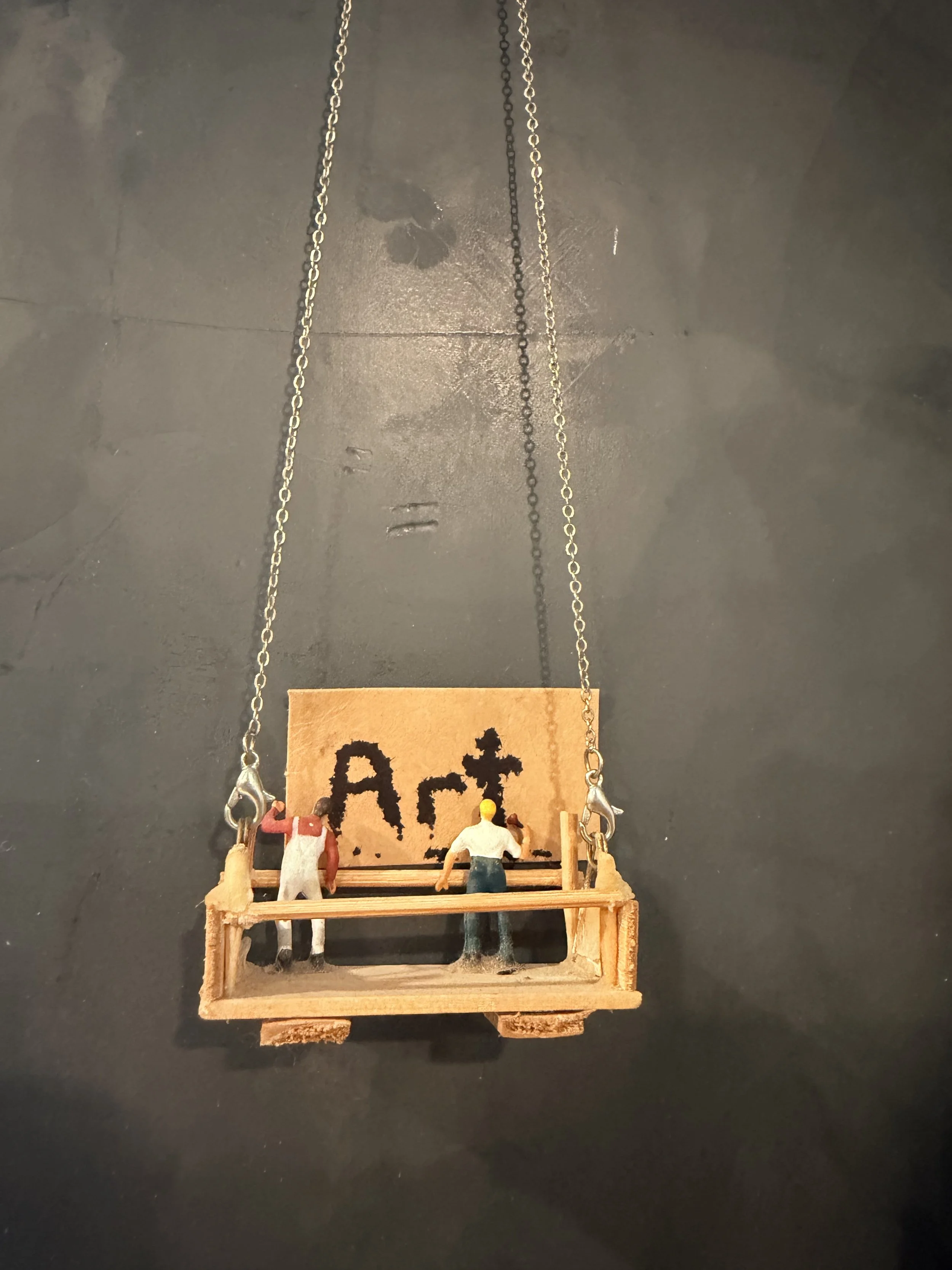 Miniature wooden swing with three small figurines of people sitting and standing inside it, set against a black background with a sign reading 'Art' hanging behind the swing.