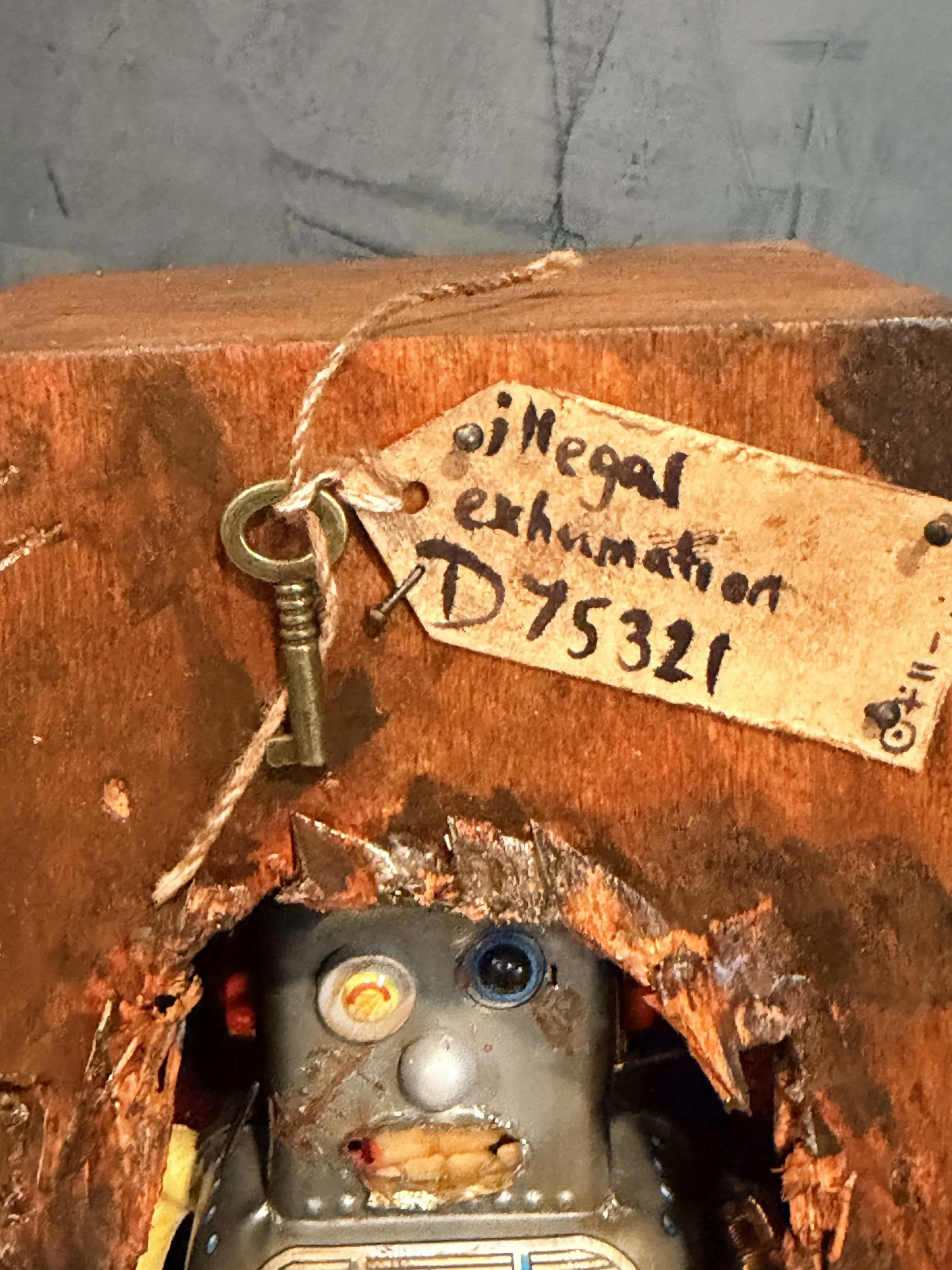Close-up of a rusted, corroded metal surface with a handwritten tag that reads 'Neilson exhaust D753321'. There is also a small key attached to the tag with string, and various mechanical or electronic components and wires underneath.