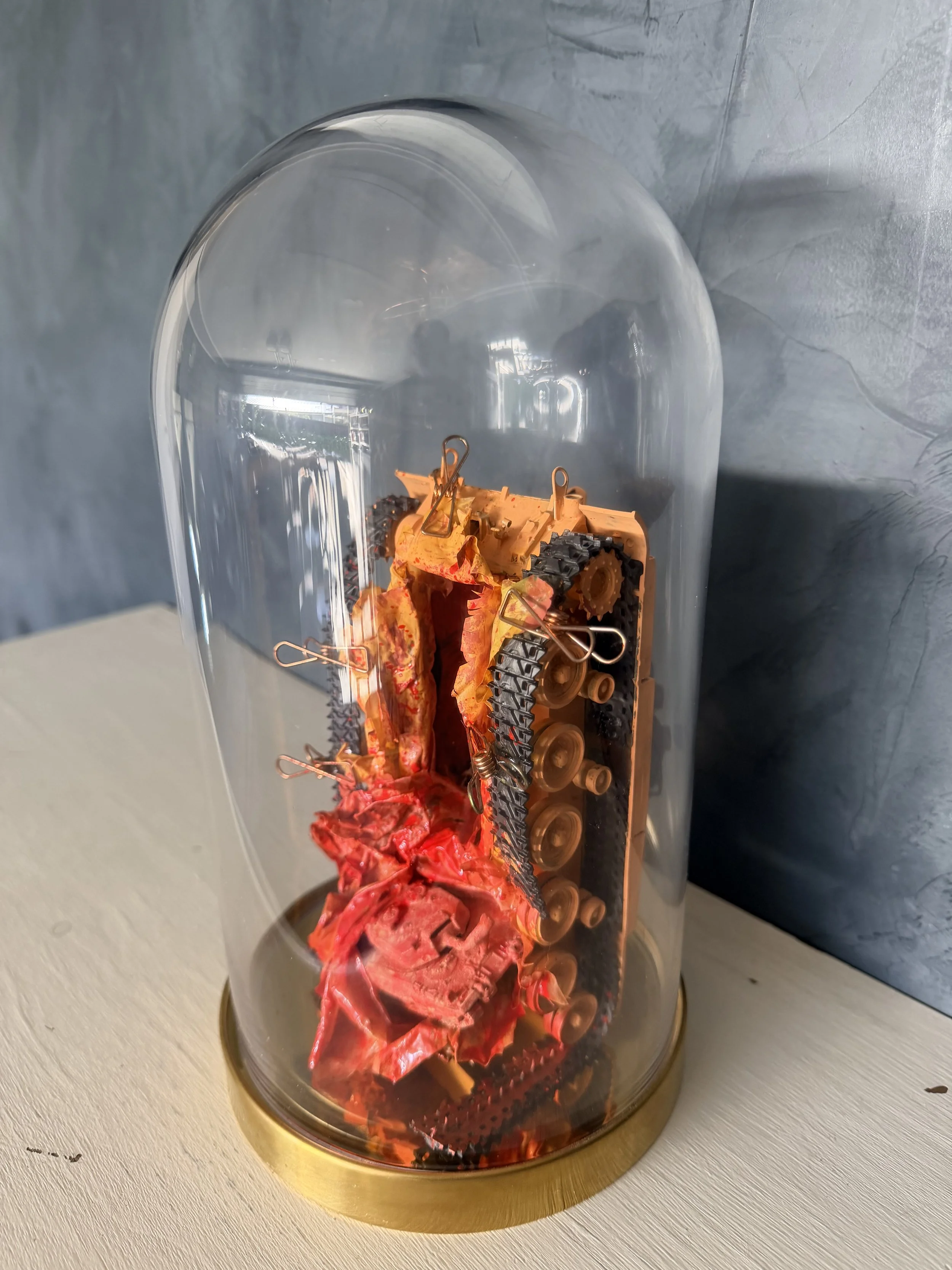 A glass dome encases a small orange toy model tracked vehicle partially covered in red tissue paper, sitting on a gold-colored base.