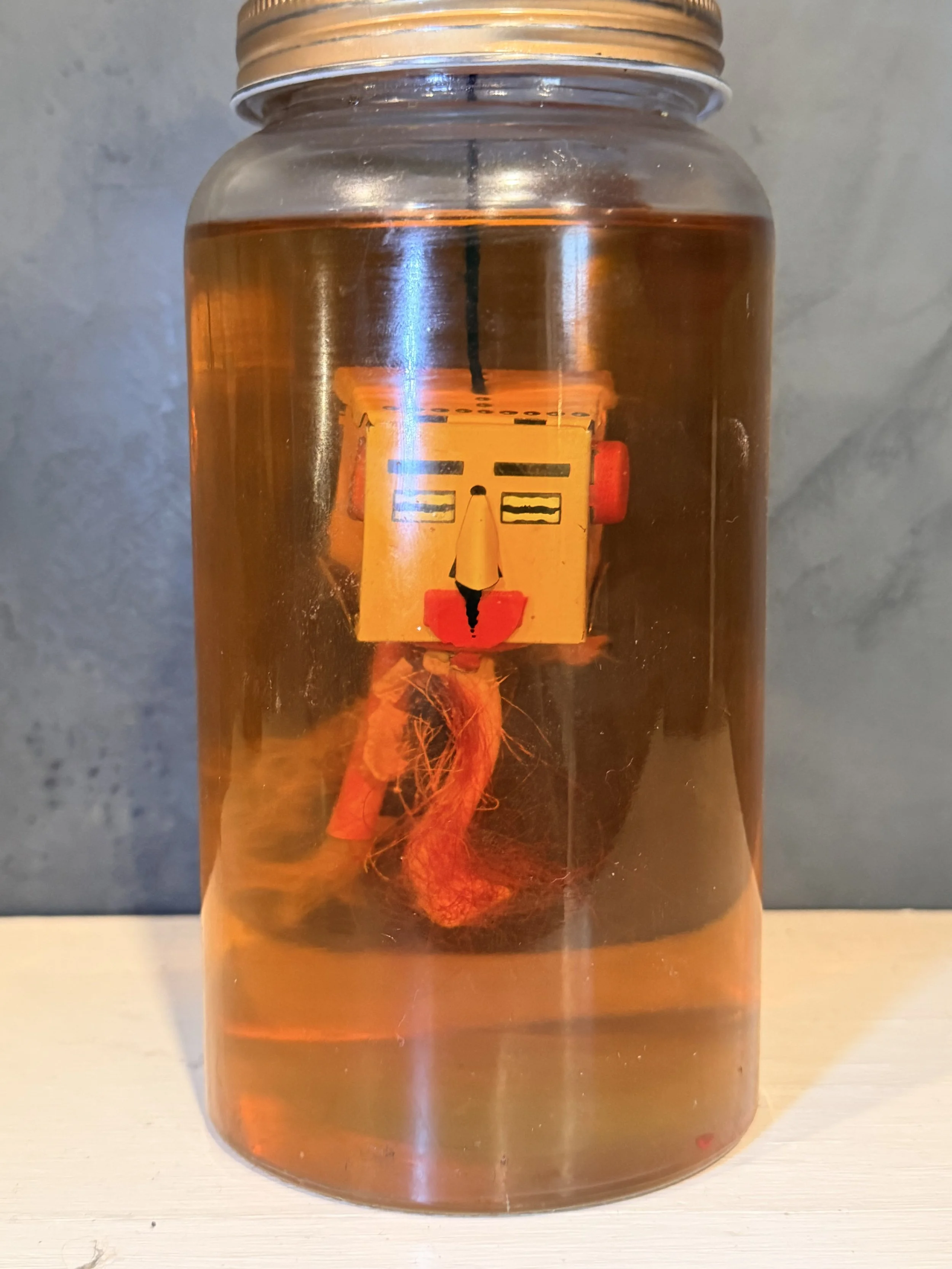 Close-up of a glass jar filled with amber-colored liquid, with a yellow and orange item inside that appears to be a piece of fabric or a cloth. The jar is placed on a light-colored surface, and the background is a plain wall.