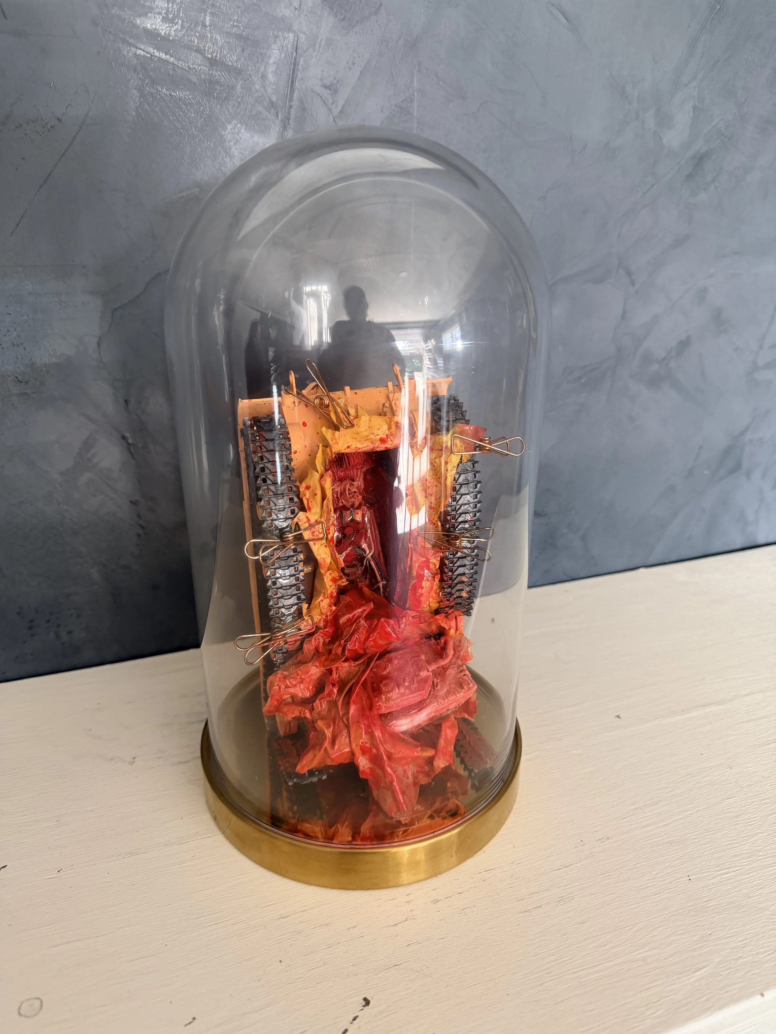 A glass dome display case with a gold-colored base showing a model of a cooking flame with crumpled orange and red paper inside, simulating fire.