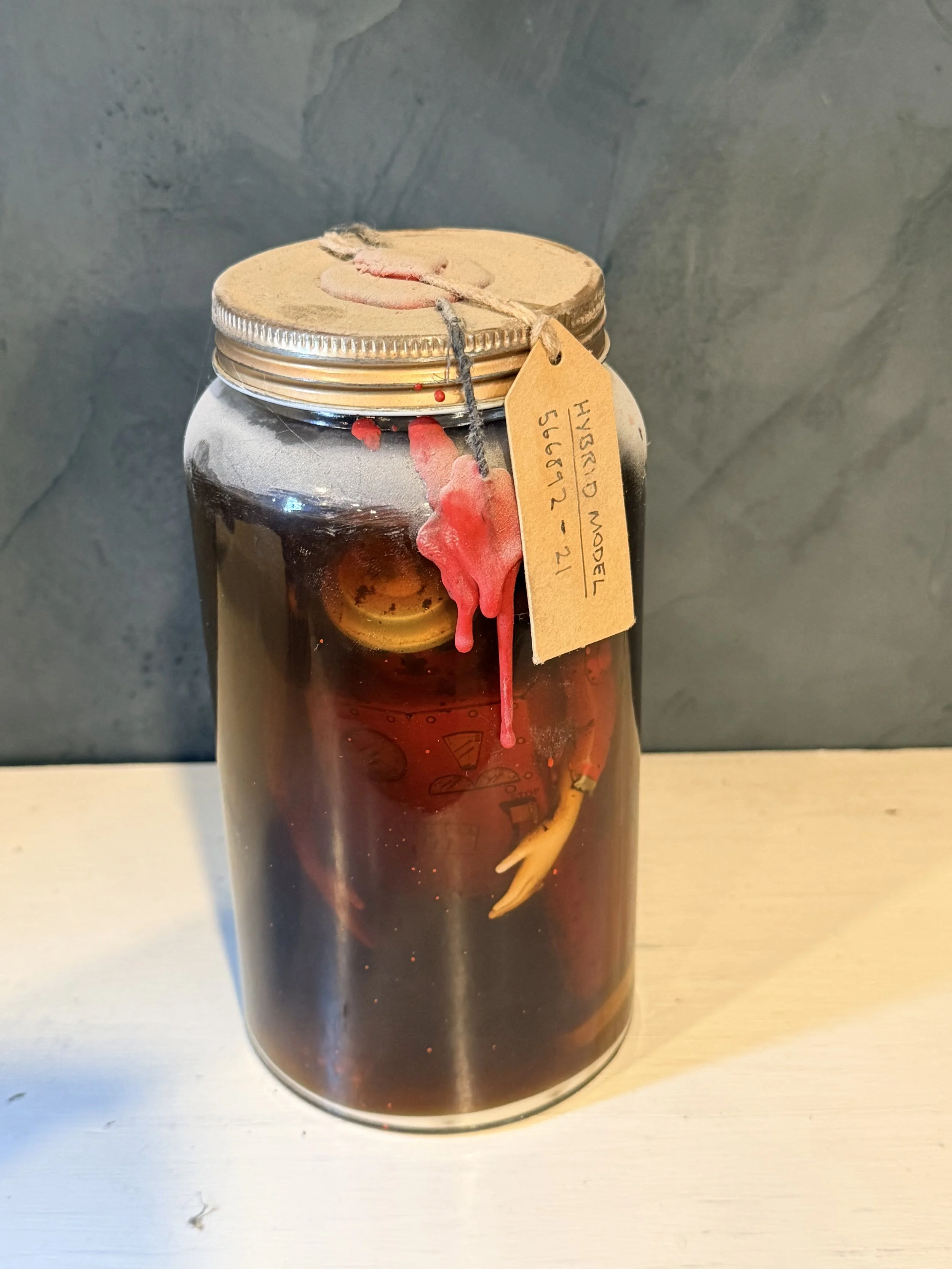 A glass jar filled with dark liquid, a string with a tag attached around its neck, a red and yellow object inside, and a pinkish wax or substance dripping down the side of the jar.