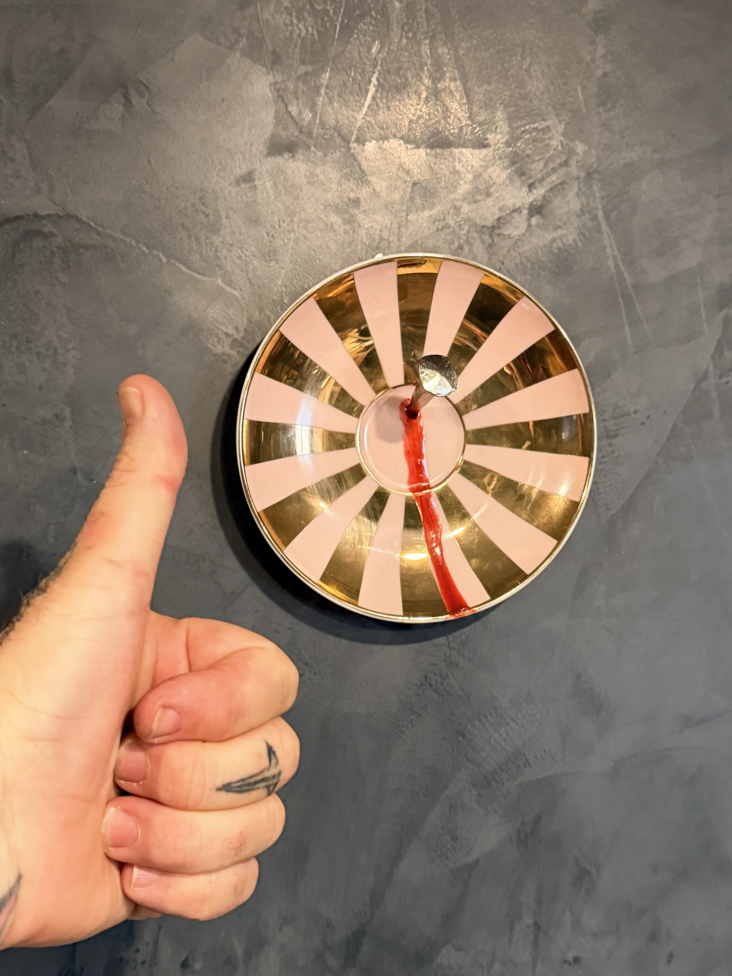 A person's hand holding a roulette wheel with pink and gold segments, with red liquid spilling from its center onto a dark surface.