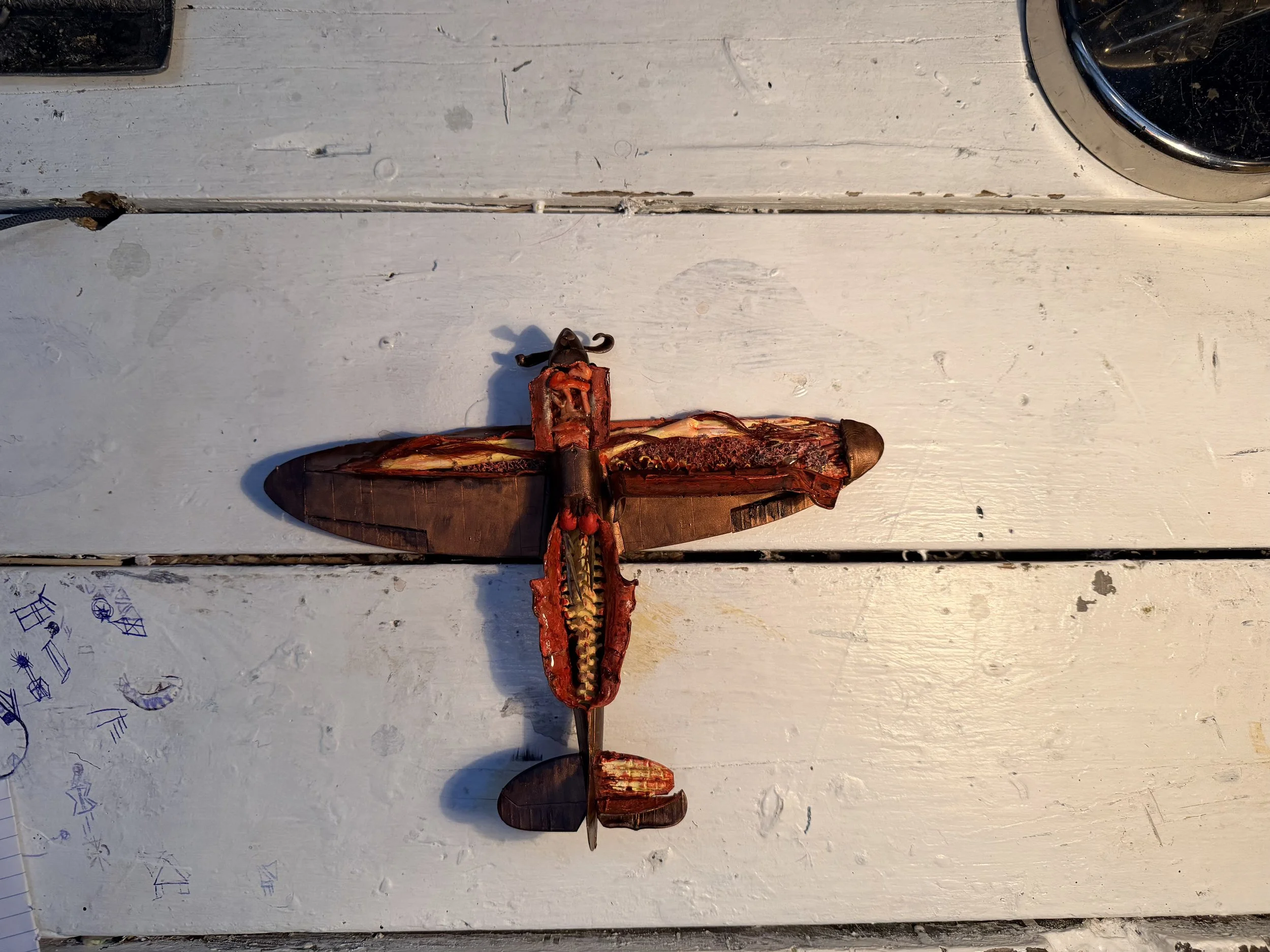 A broken butterfly-shaped decorative item with reddish-brown color, lying on a white wooden surface with visible scratches, stains, and random blue ink sketches.