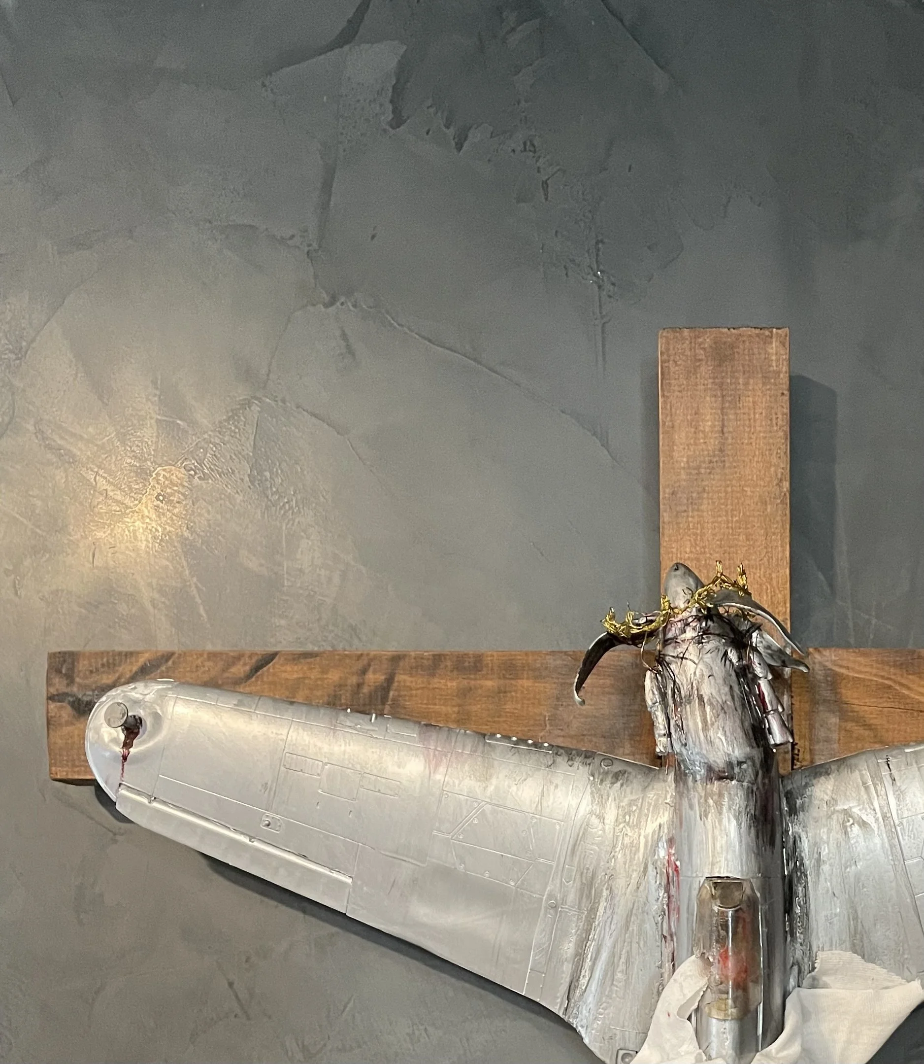 A silver metallic sculpture of a fish mounted on a wooden plank, with some fabric and decorative elements at the bottom, situated against a dark gray wall.