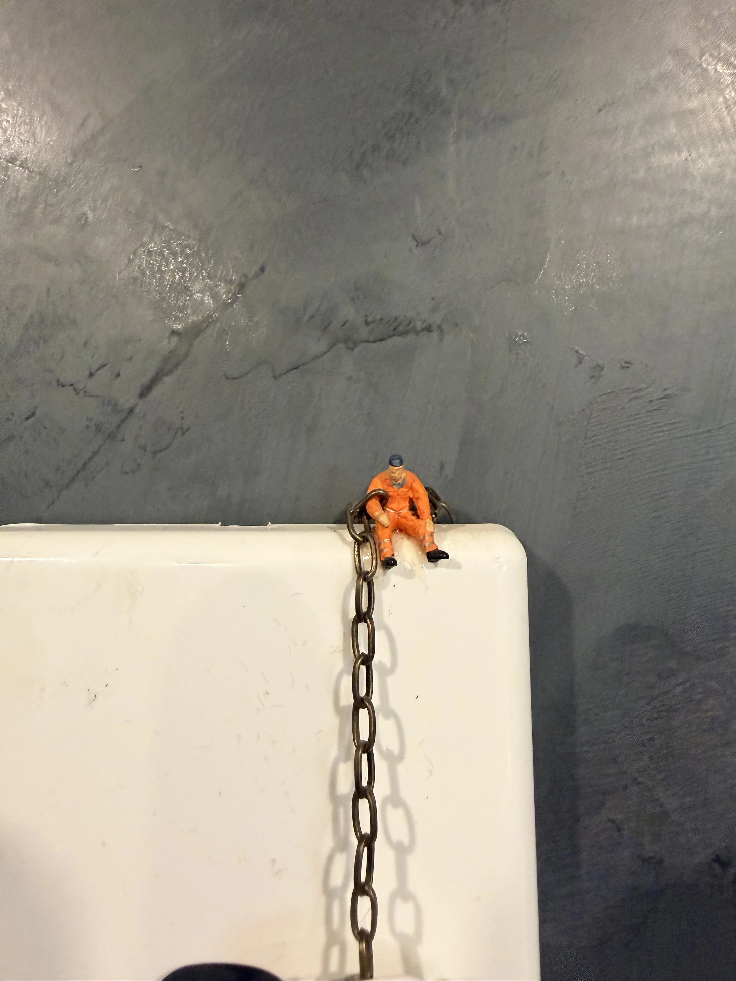 A miniature figurine of a person wearing an orange jumpsuit and a grey cap, sitting on a white surface, with a chain hanging down from above.