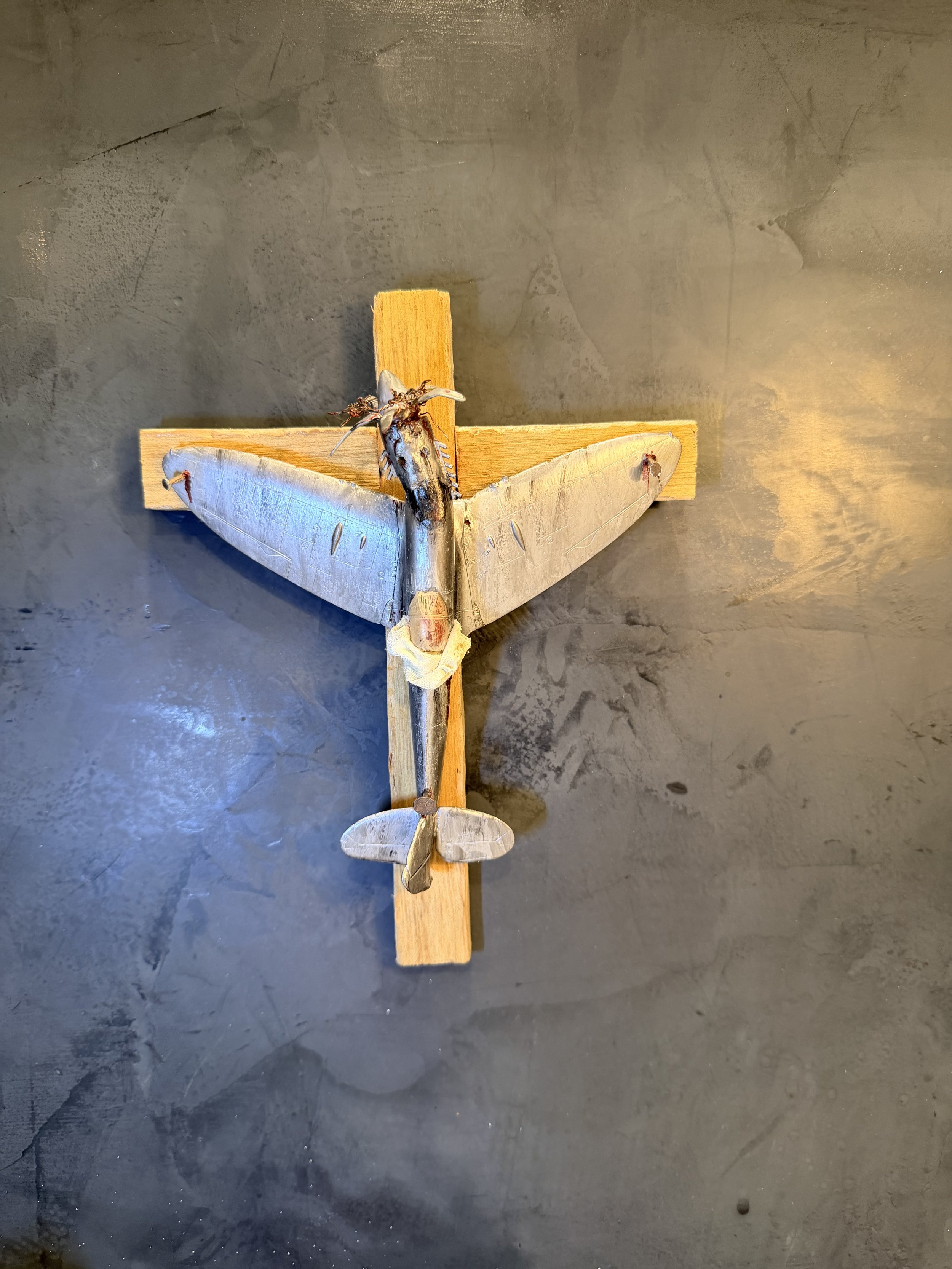 A model or toy airplane mounted on a wooden cross with a tiny creature, possibly a bug or spider, on the top wing.