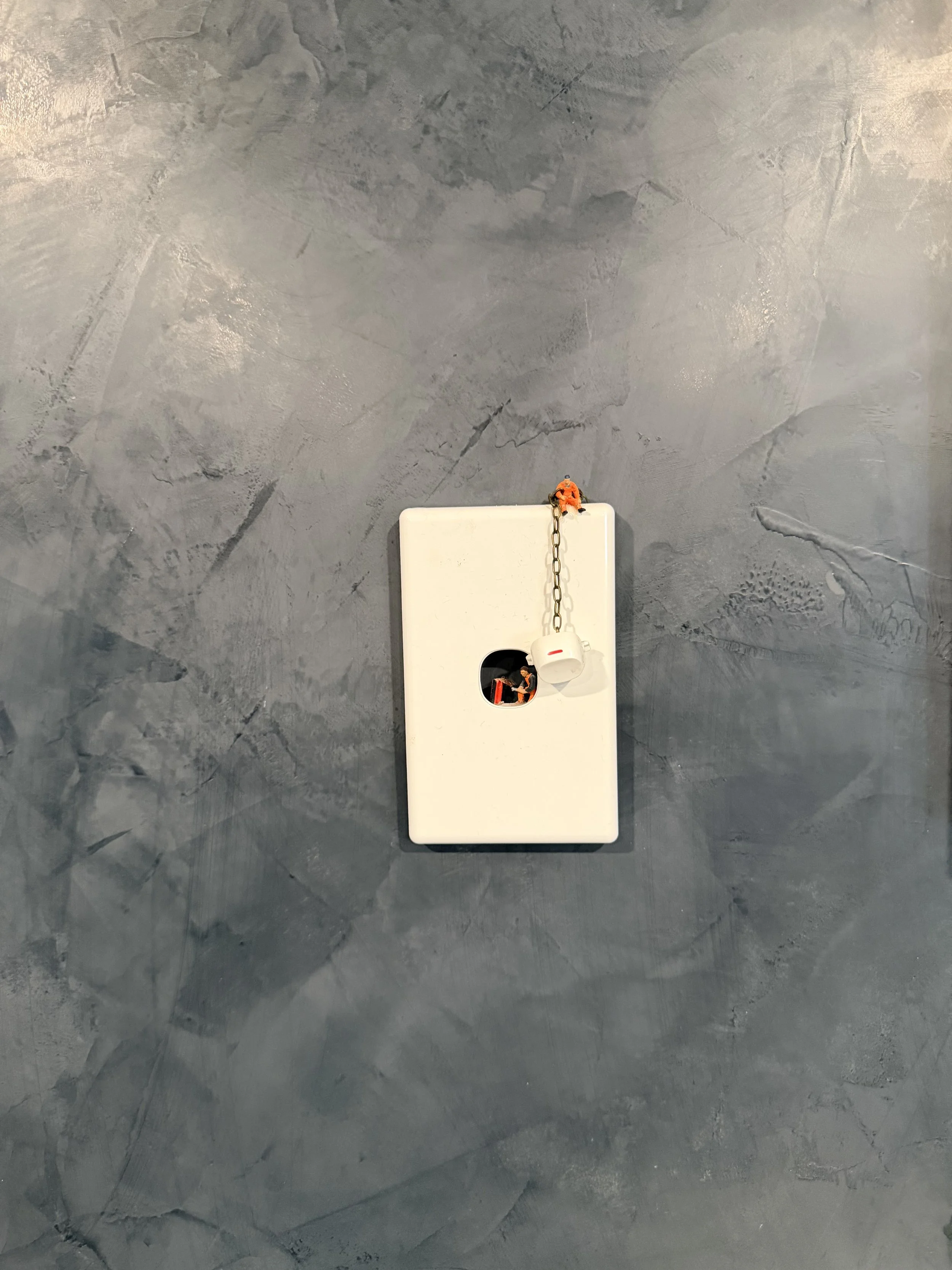 A gray textured wall with a white plastic electrical panel in the center. Two miniature figurines of construction workers are positioned on the panel, one on top and one inside a hole, both holding tools. A chain is hanging from the top of the panel.