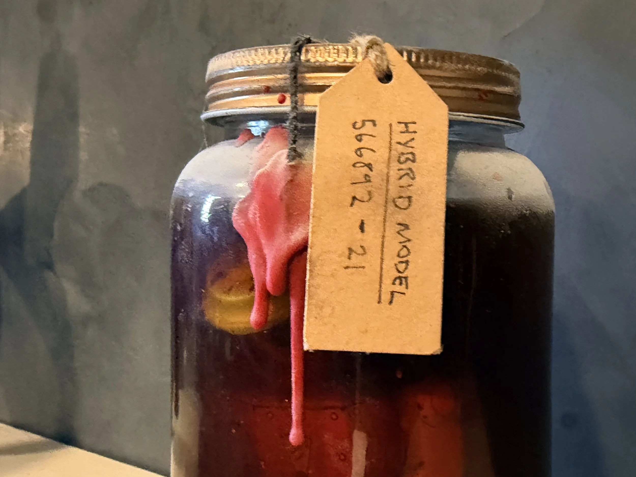 A glass jar secured with a black rubber band, containing pink wax with a wick, and tagged with a yellow label that has handwritten text, on a metallic surface.