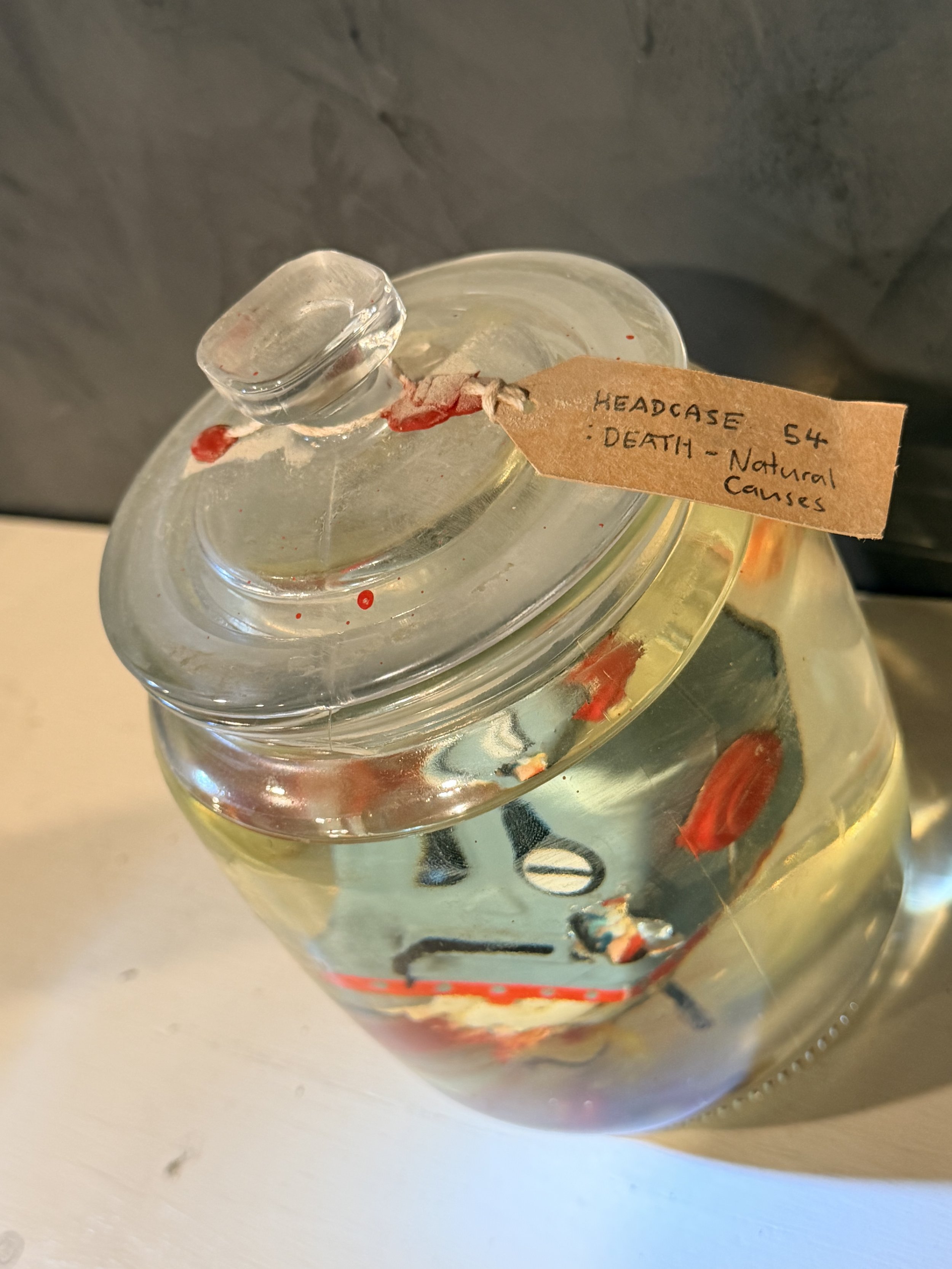 A clear glass jar filled with water and small red fish swimming inside. The jar has a paper tag attached with string, labeled 'HEADCASE 54 : DEATH - Natural Causes'.