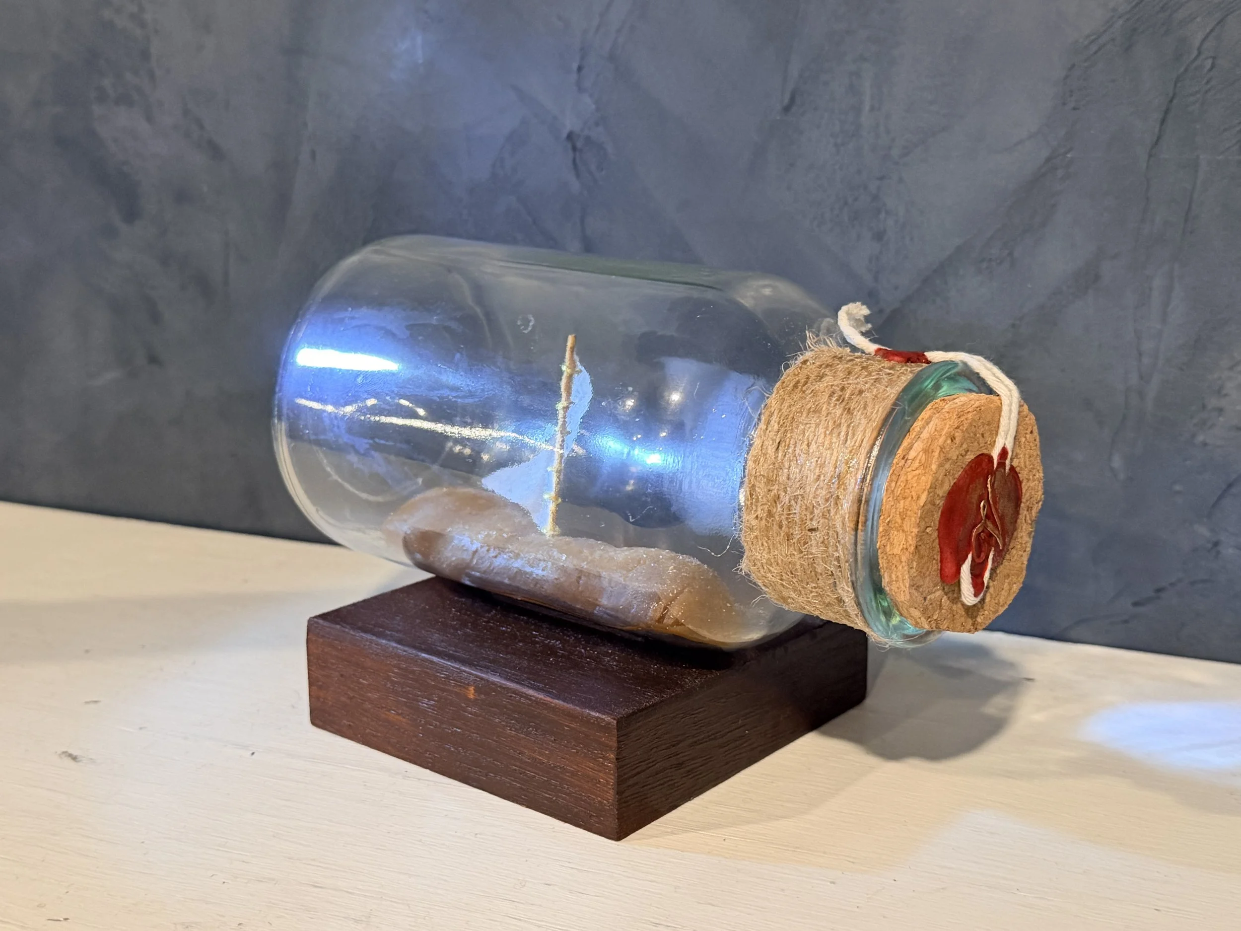 A small glass container featuring a cork stopper wrapped with red and white string, mounted on a dark wooden base. The container has a visible light bulb filament inside, lit to glow with a blue hue, creating a decorative or artistic display.