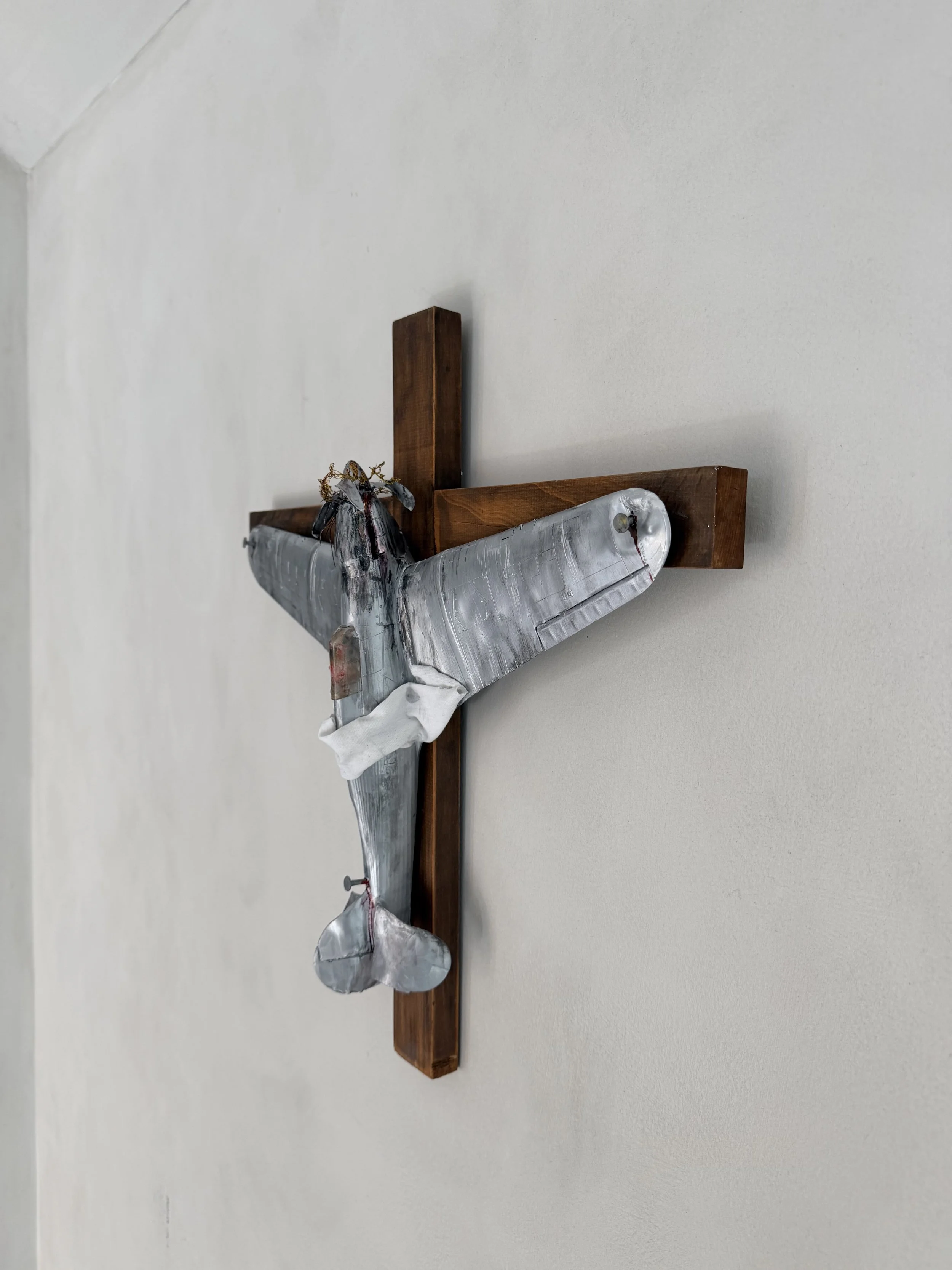 A metal whale sculpture mounted on a wooden cross against a plain white wall.