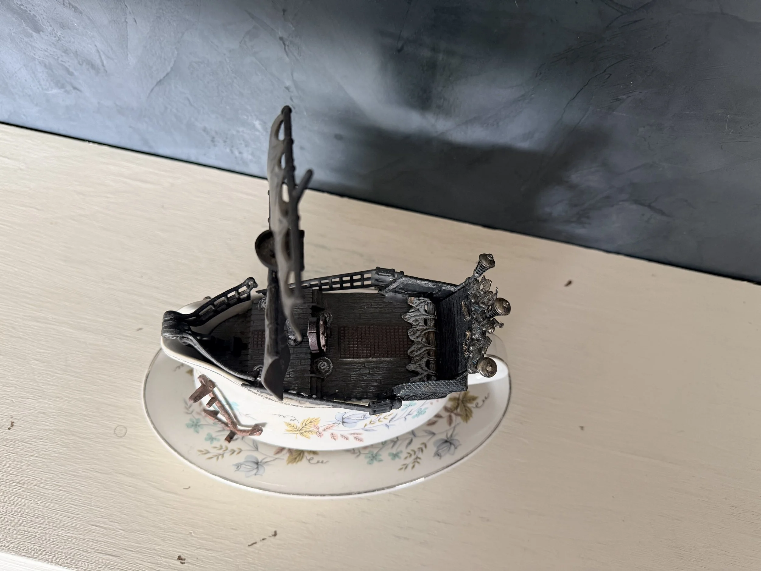 A decorative miniature castle with black walls and silver accents, placed inside a porcelain cup with floral patterns, on a white surface with a blue textured wall in the background.