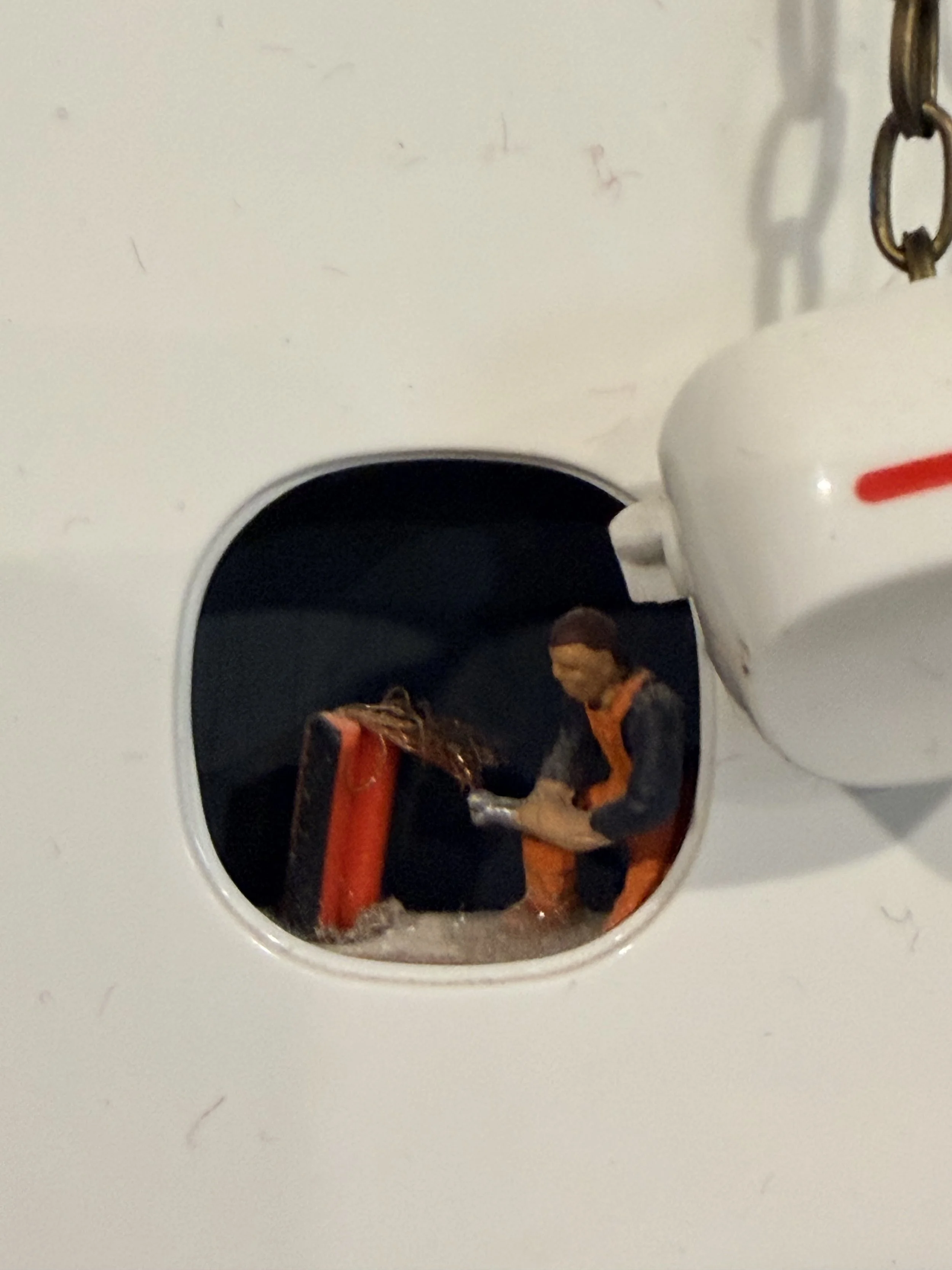 A person wearing an orange jacket is seen through a circular opening in a white surface, with a partially visible white appliance with a red marking nearby.