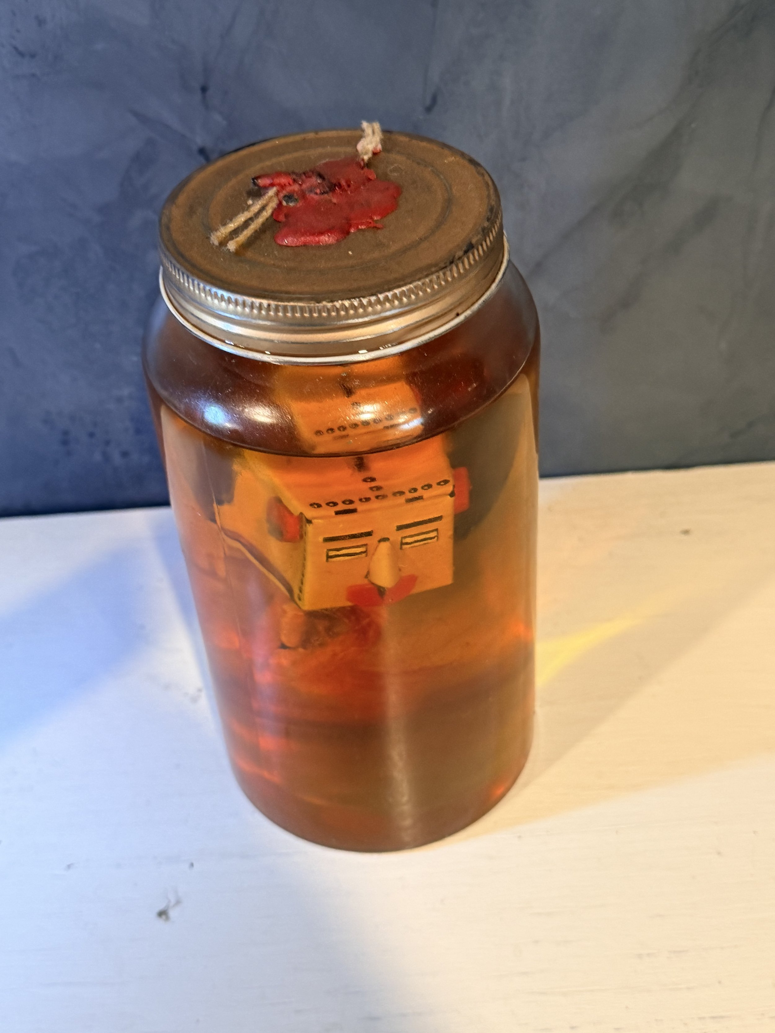 A glass jar filled with a dark amber liquid, sealed with a metal lid that has some red wax and string on top, is placed on a light-colored wooden surface against a gray background.