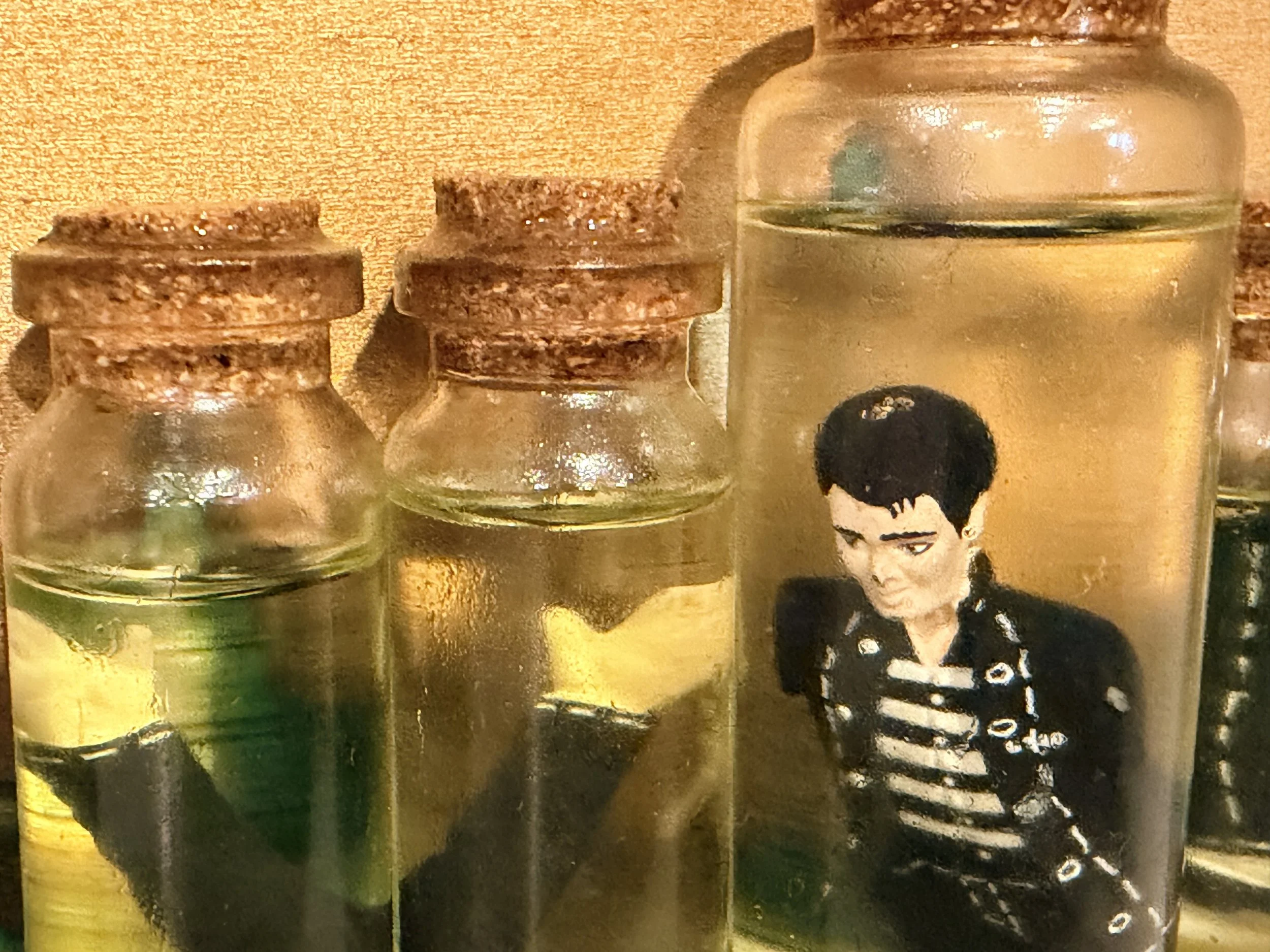 Close-up photograph of small glass jars with cork lids, containing various powders or substances, placed on a textured surface. Part of a person's portrait with black hair and a striped top is also visible among the jars.