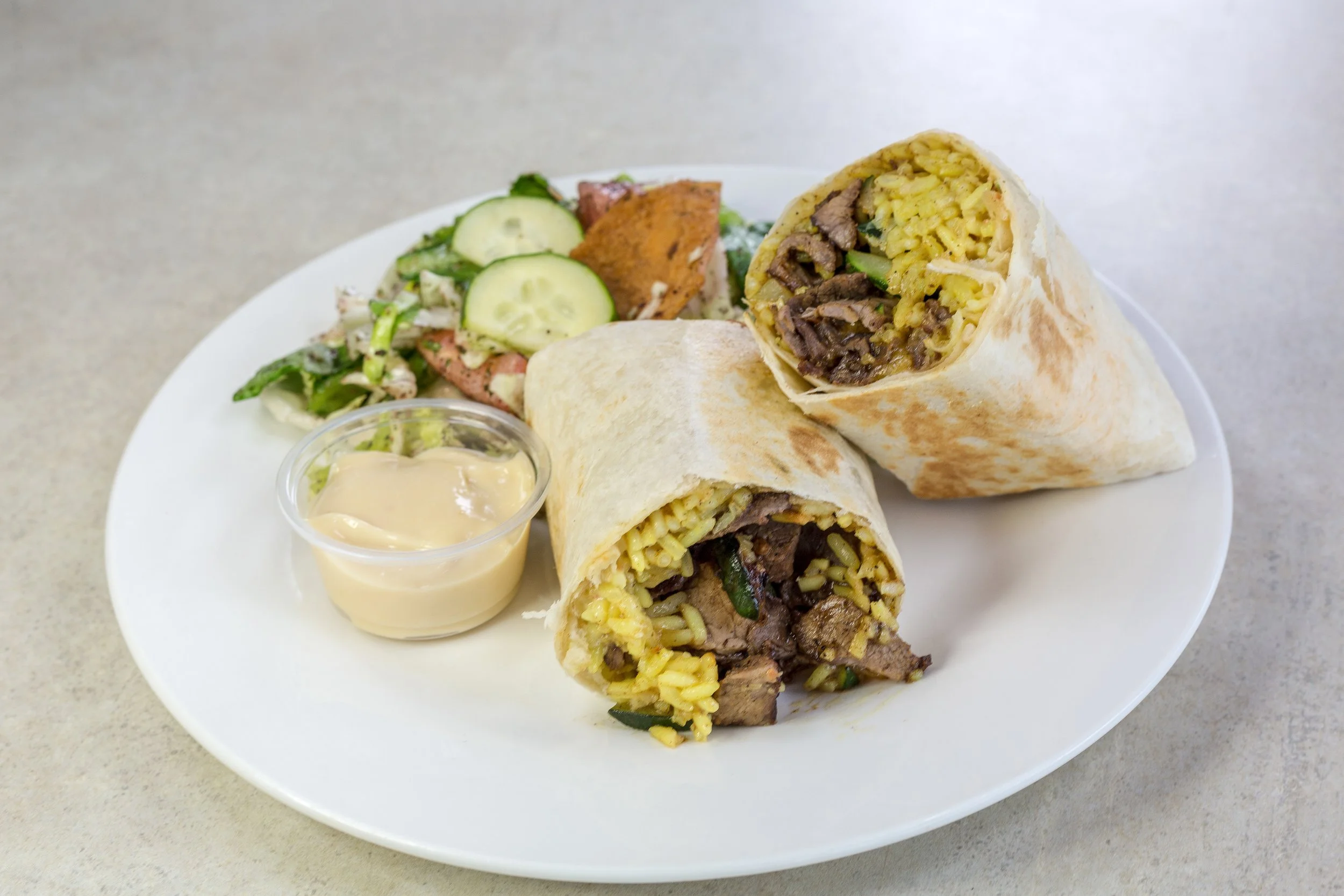 Two beef and rice burritos cut in half on a white plate, with a side salad of cucumbers and lettuce, and a small cup of creamy sauce.
