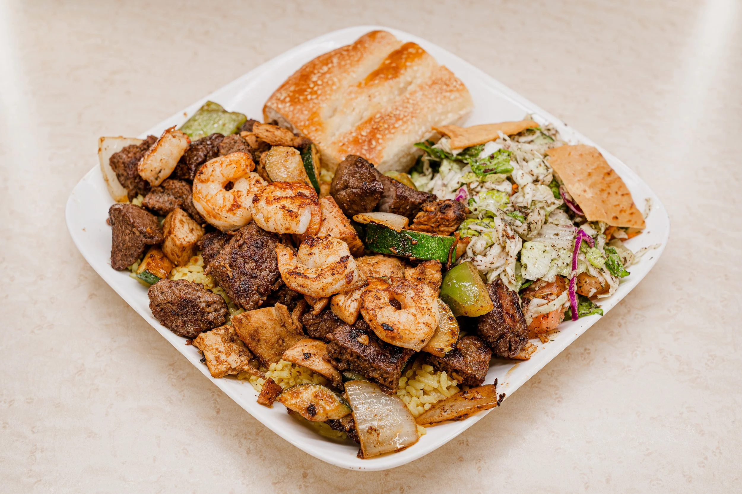 A white plate filled with grilled meats, shrimp, vegetables, rice, a piece of garlic bread, and a side salad.