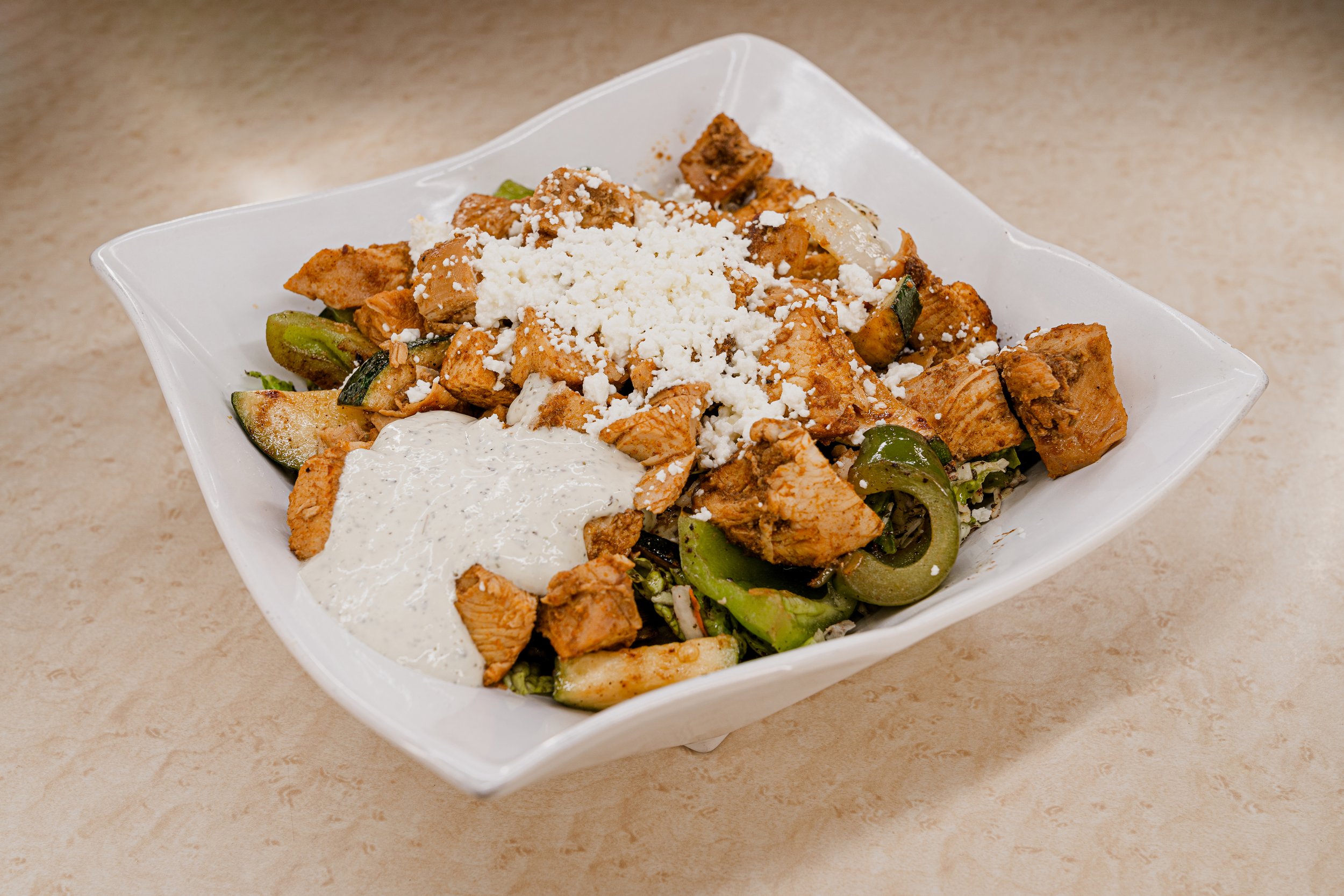 A white bowl filled with a Mexican dish containing grilled chicken, zucchini, green peppers, topped with crumbled cheese and sour cream.
