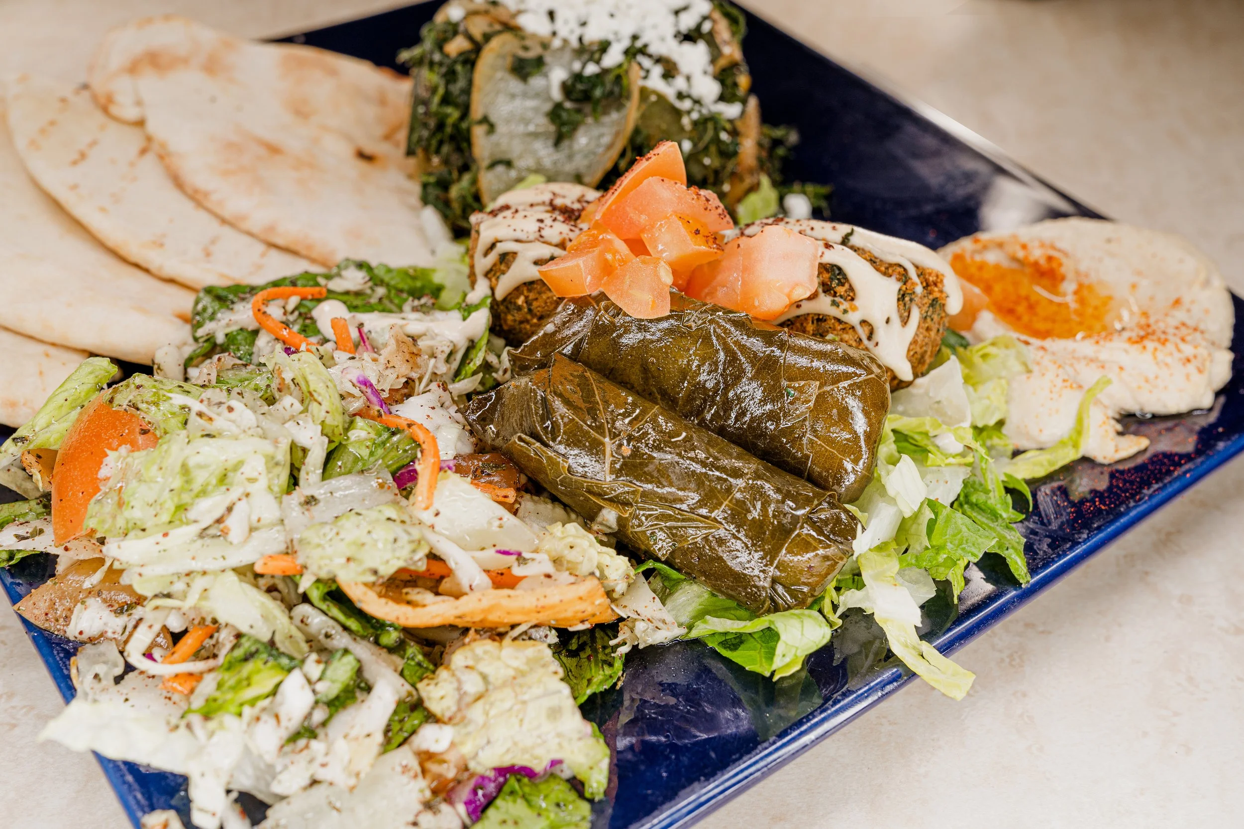 A blue rectangular plate holding various Mexican foods, including tacos, salad, stuffed grape leaves, and dips, with a beige table surface in the background.