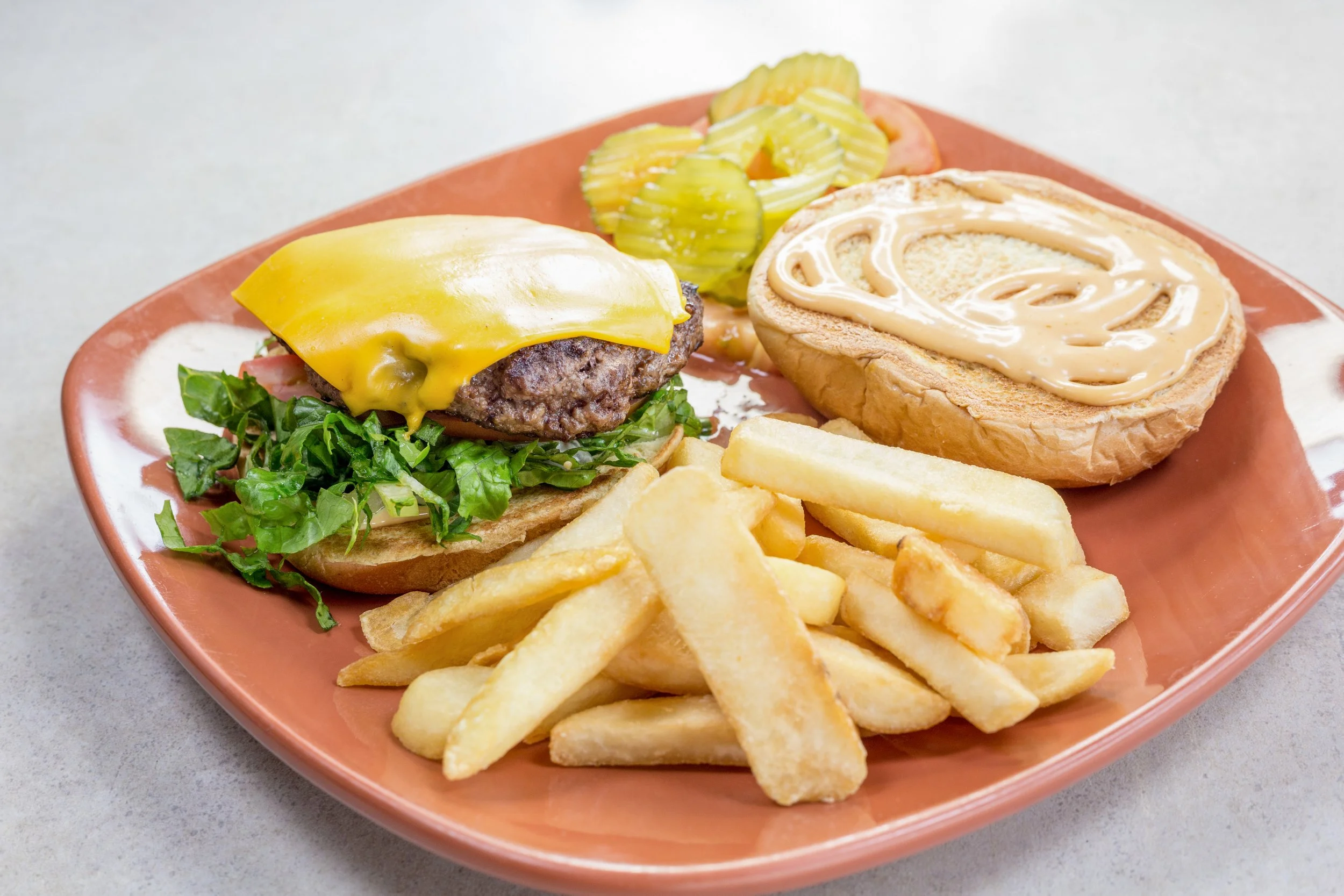 A cheeseburger in a bun with cheese, lettuce, and a beef patty, served with crinkle-cut fries and pickles on a pink plate.