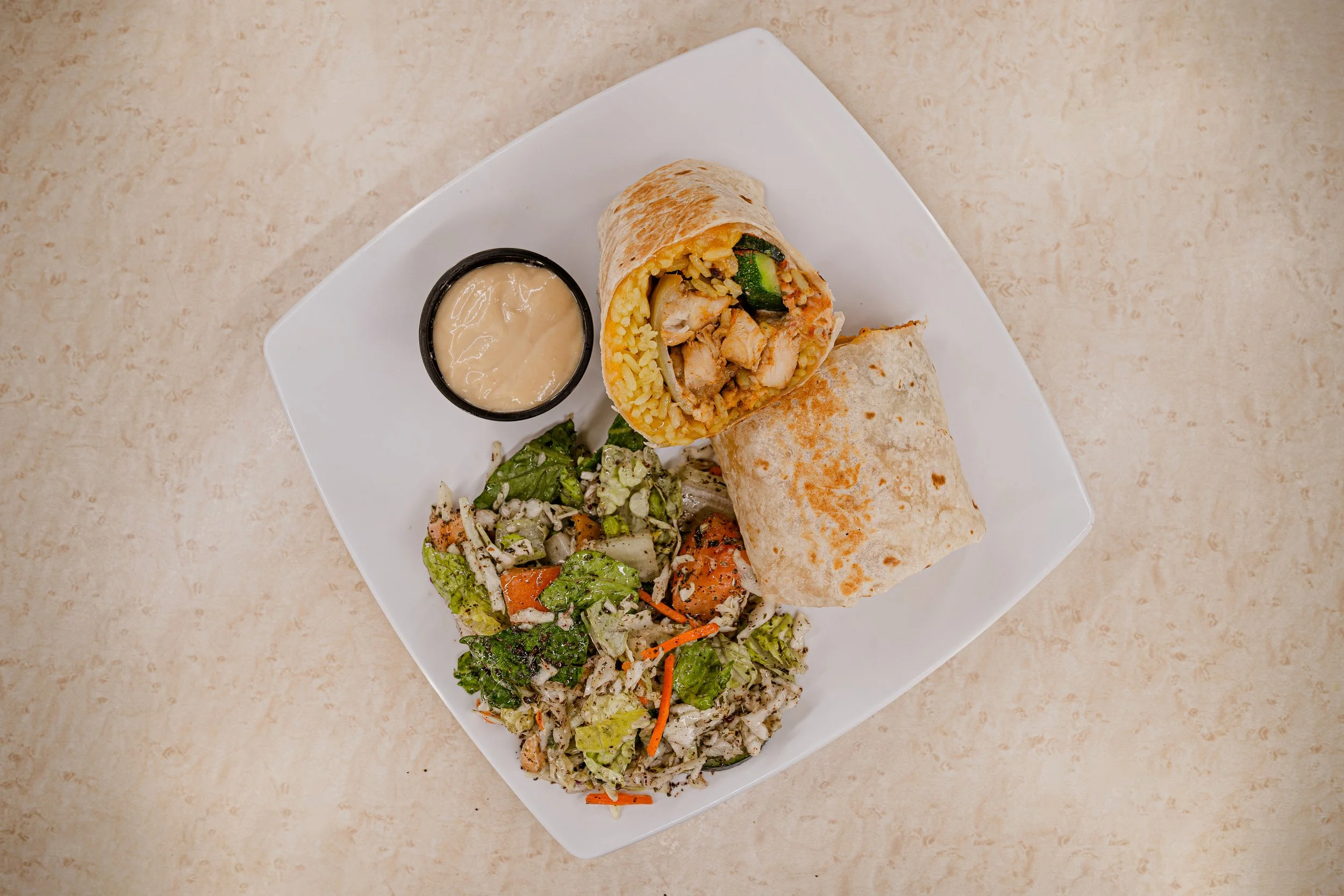 A plate with a burrito cut in half, a side salad, and a small container of sauce on a light-colored table.