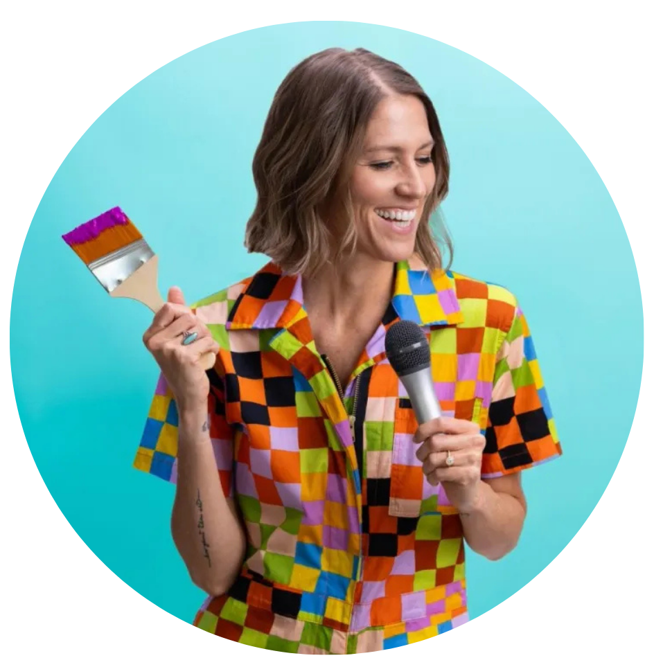 A woman with shoulder-length brown hair holding a small paintbrush with purple paint and a microphone, smiling against a light blue background.
