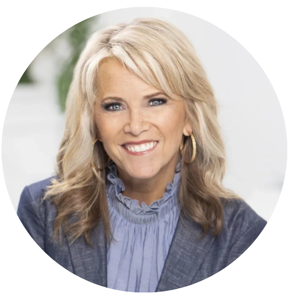 A smiling blonde woman with wavy hair wearing a blue blouse with ruffled collar and a gray blazer, posing indoors.