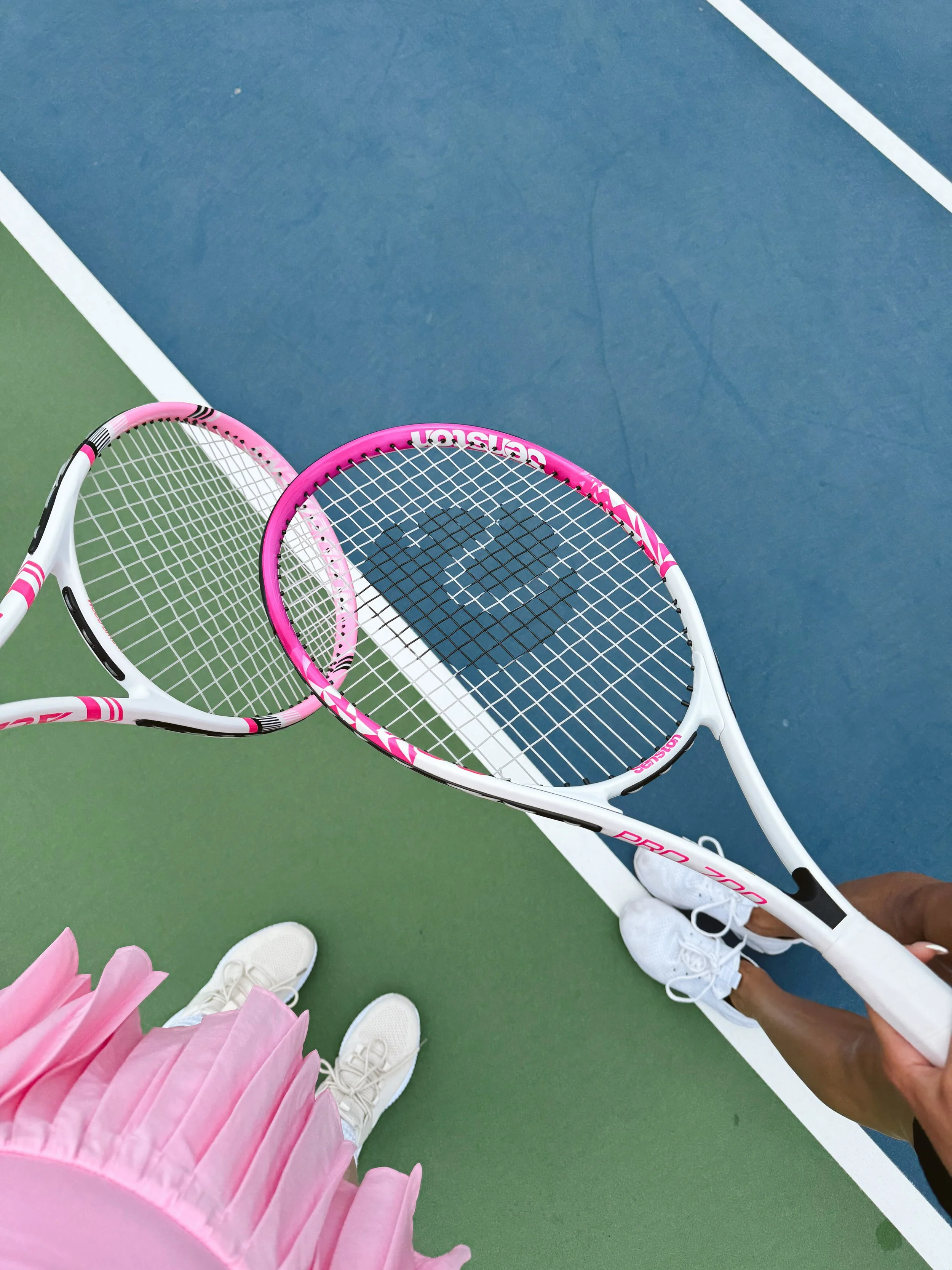 Two tennis rackets, one pink and one white, held by a person on a tennis court with a green and blue surface; also visible are pairs of white shoes, a pink flower decoration, and a person wearing white sneakers.