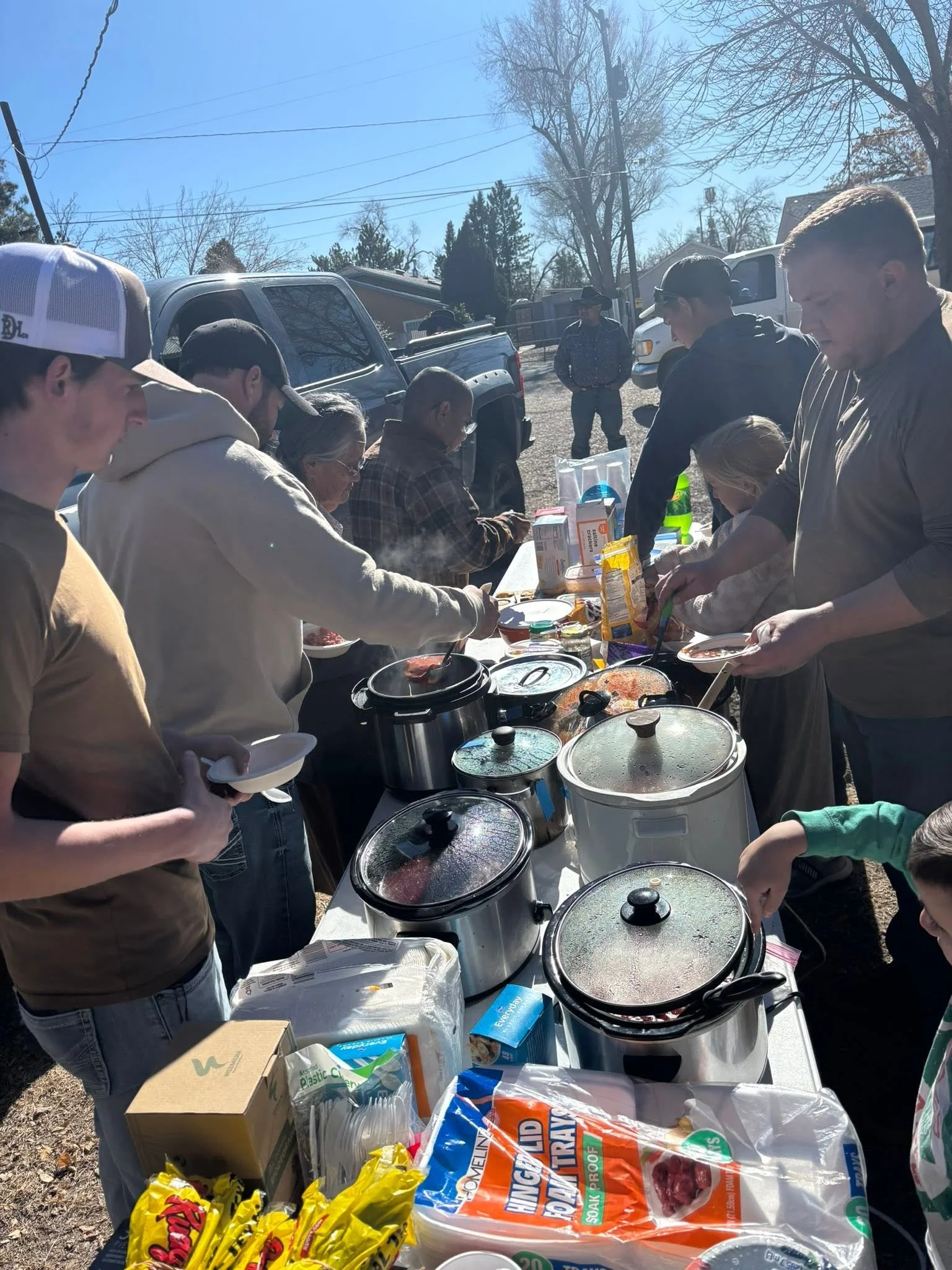 People gathered outdoors around a table with food and supplies, serving and preparing meals on a sunny day with leafless trees and parked cars in the background.