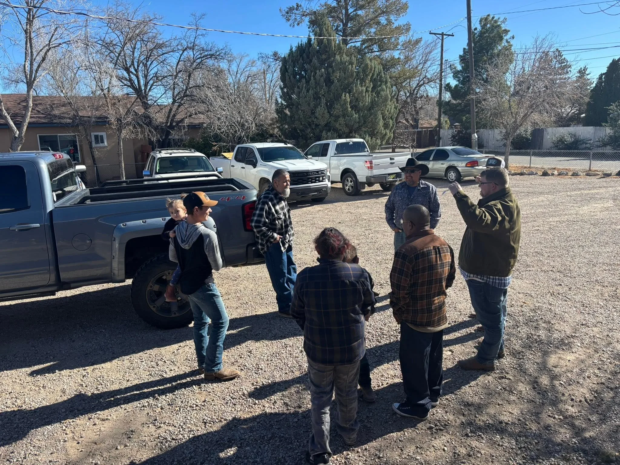 A group of people standing and talking outdoors in a gravel lot with several parked trucks and cars behind them. There are leafless trees and a house with a shingled roof under a clear blue sky.
