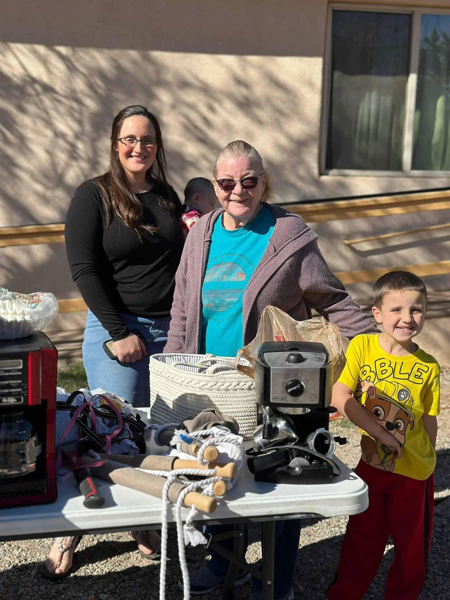 Group of people at an outdoor yard sale with various items on a table, including kitchen appliances, tools, and household items, with sunlight and shadows on the wall behind.