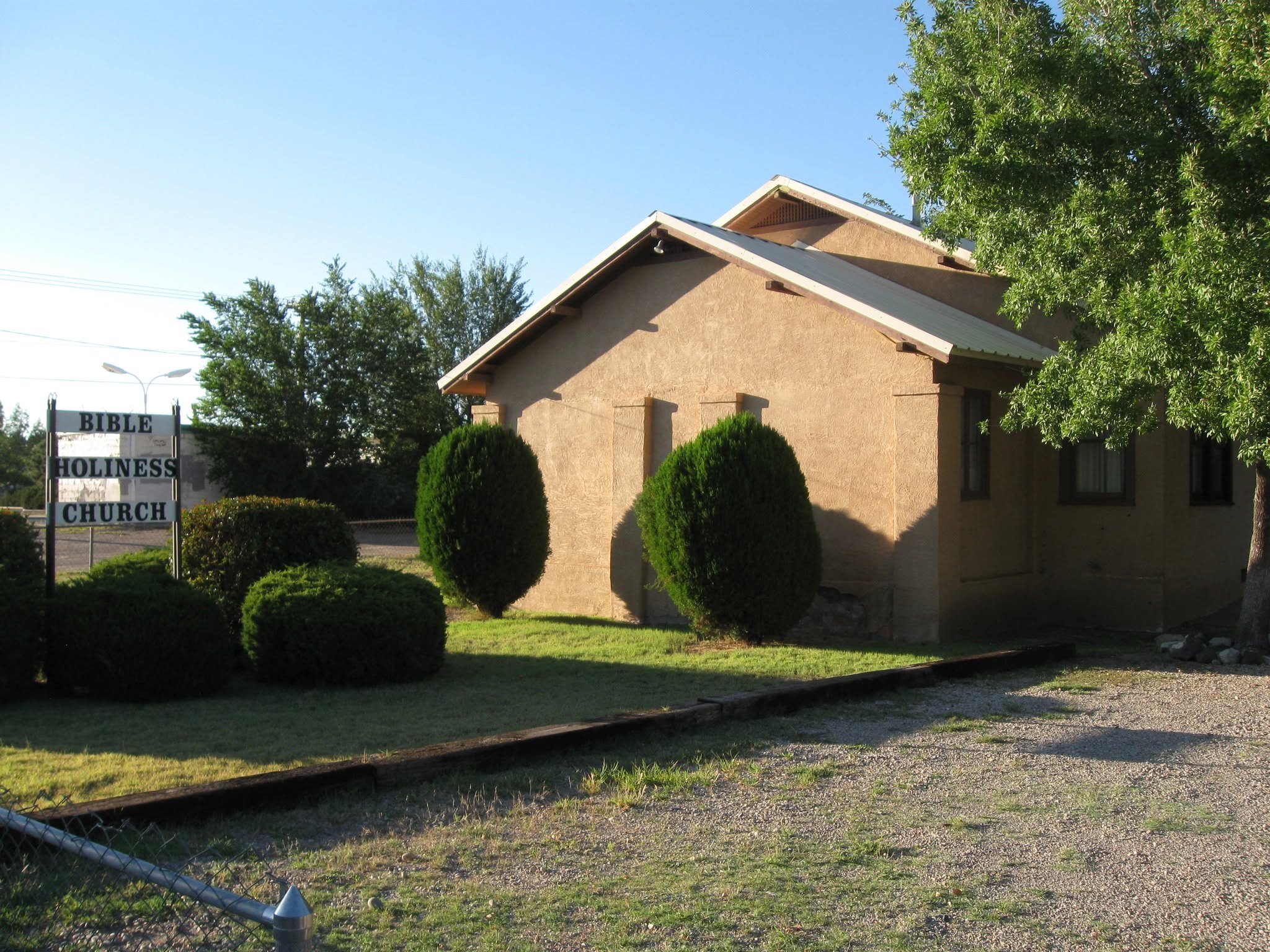 A beige church building with a sign that reads 'Bible Holiness Church' in front. The building has a sloped roof and is surrounded by green bushes and a large tree. The sky is clear and blue.