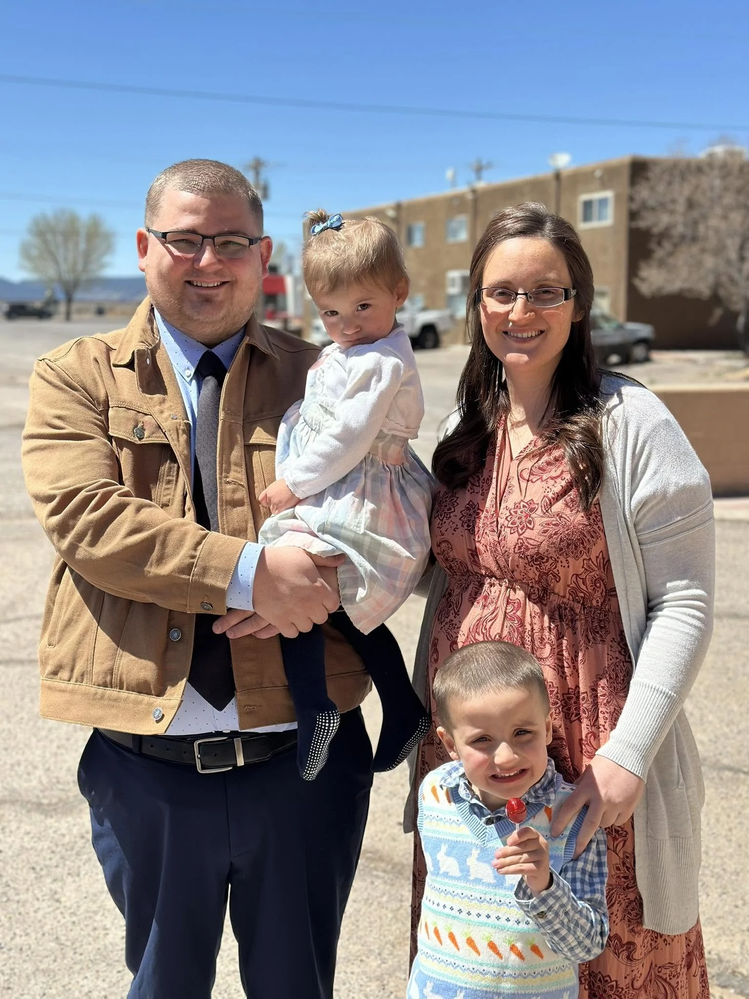 A family of four posing outdoors on a sunny day. The father is holding a young girl with a blue bow in her hair, and the mother is standing next to an older boy holding a lollipop. The background shows a clear blue sky, a building, and some trees.