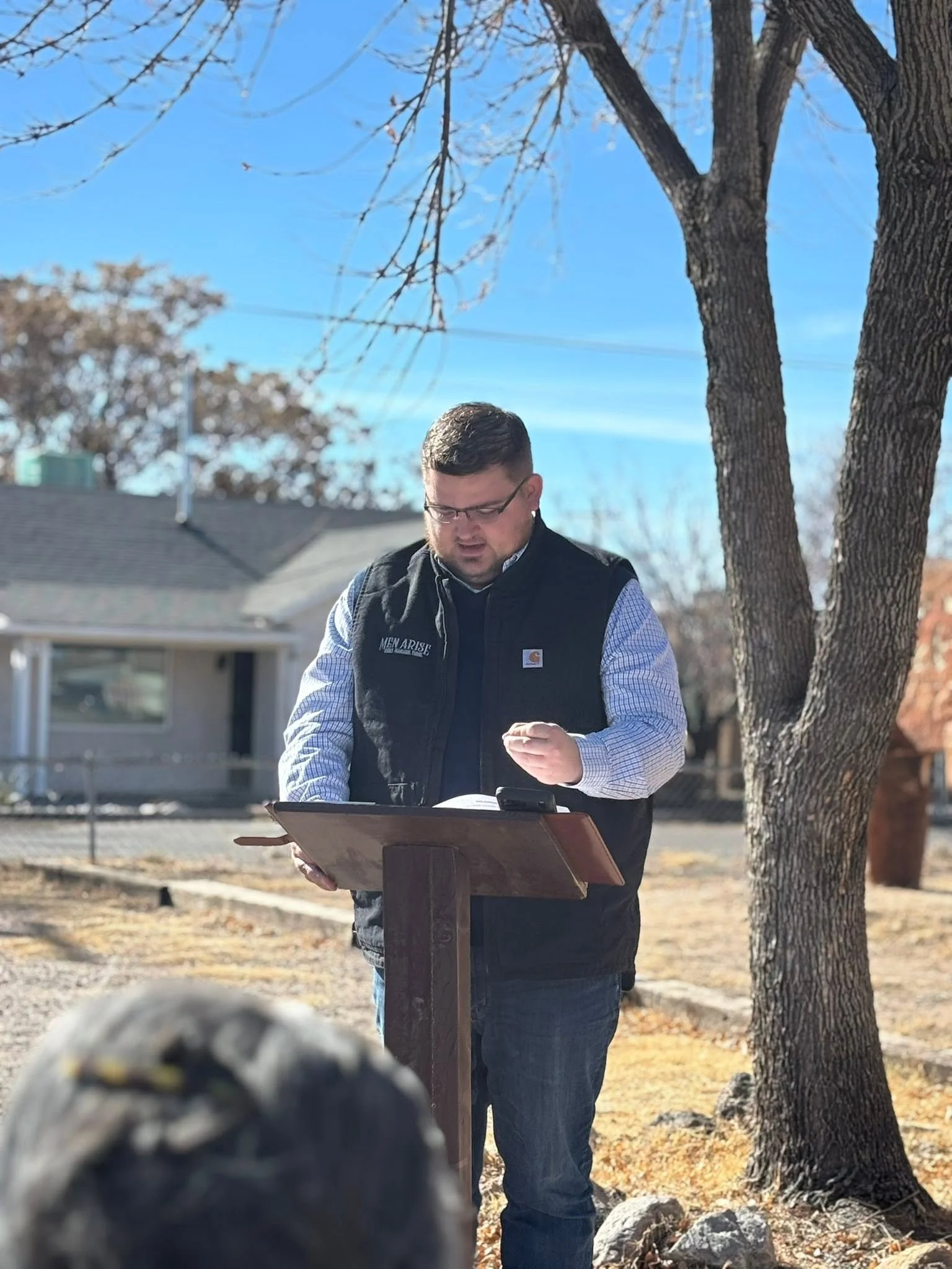 A man with glasses, wearing a black vest over a blue and white checkered shirt, stands outdoors reading from a paper on a wooden stand. There is a leafless tree behind him and houses in the background under a clear blue sky.