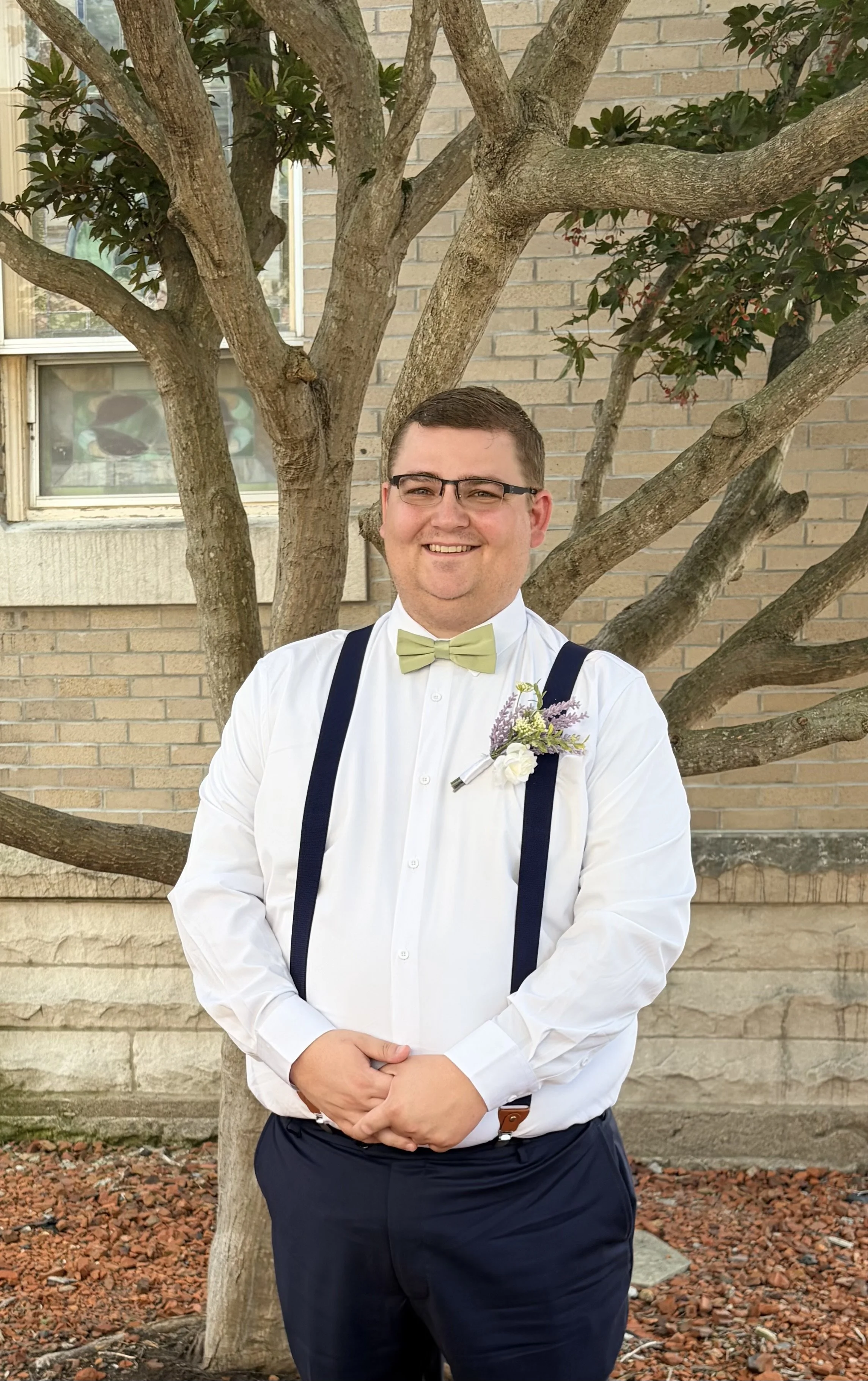 A smiling man in formal attire with a white shirt, green bow tie, and suspenders, standing outdoors in front of a tree and brick building. He has short brown hair, glasses, and is clasping his hands in front of him.
